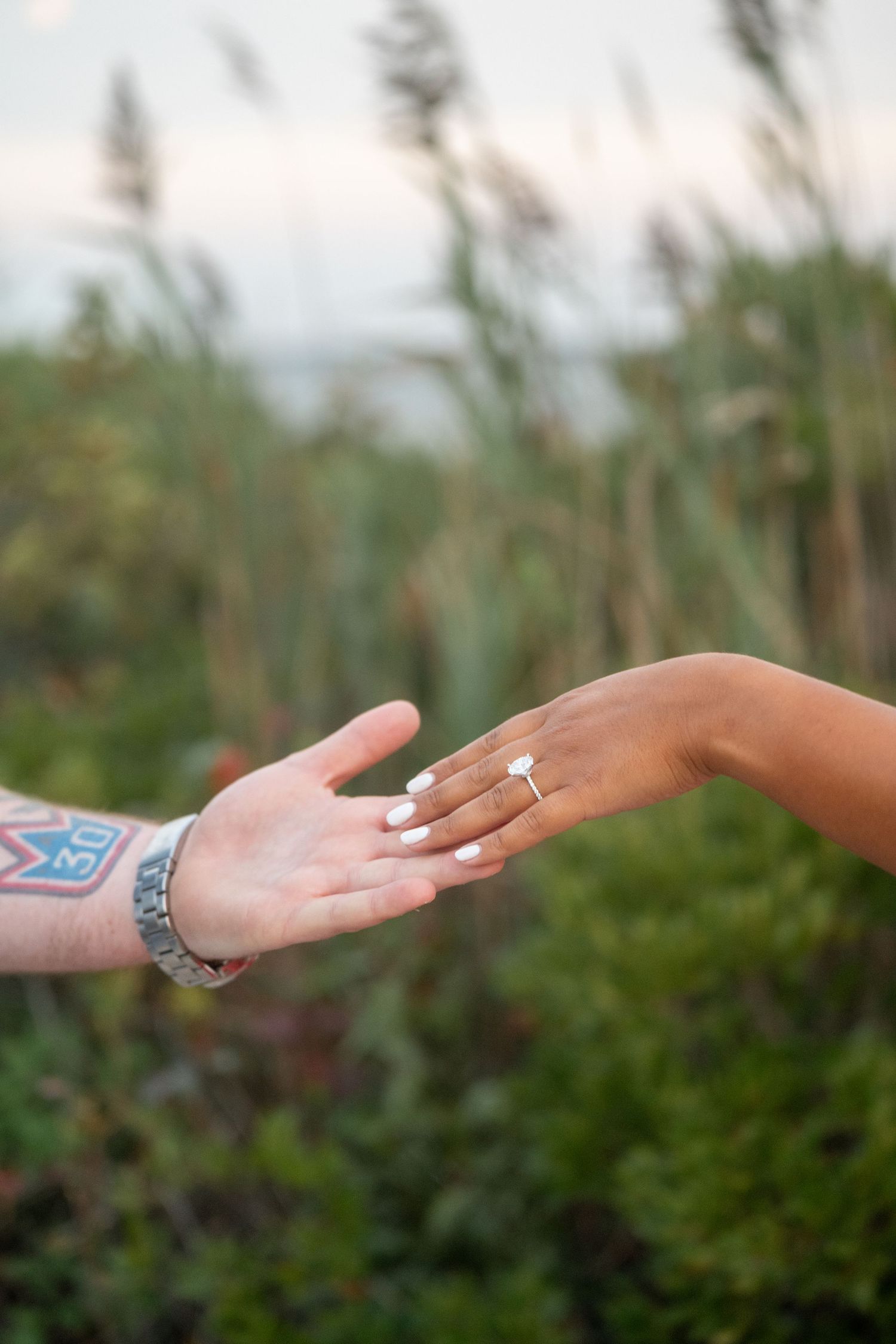 Two hands reaching towards each other with one wearing an engagement ring, set against a blurred natural background.