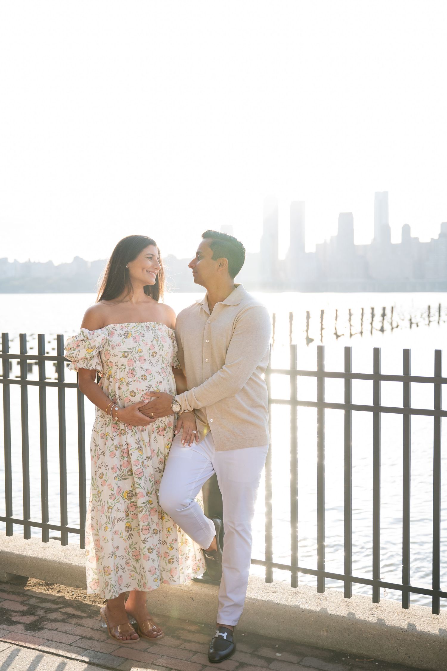 Couple in floral dress and white outfit pose together on waterfront promenade with city skyline in background.