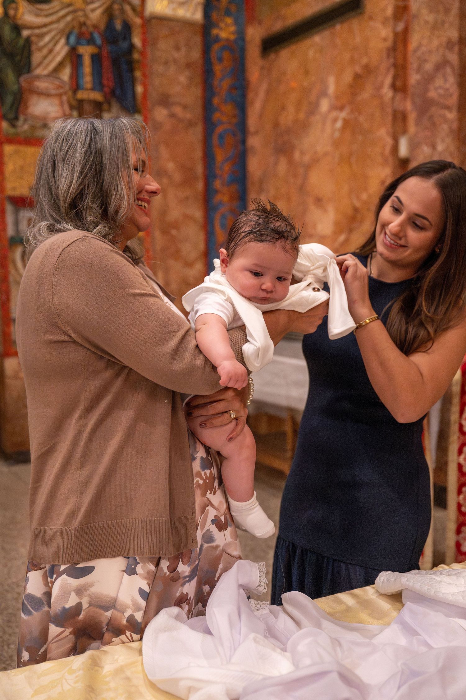 A baby in white clothing is held during a special church ceremony with decorative walls in the background.