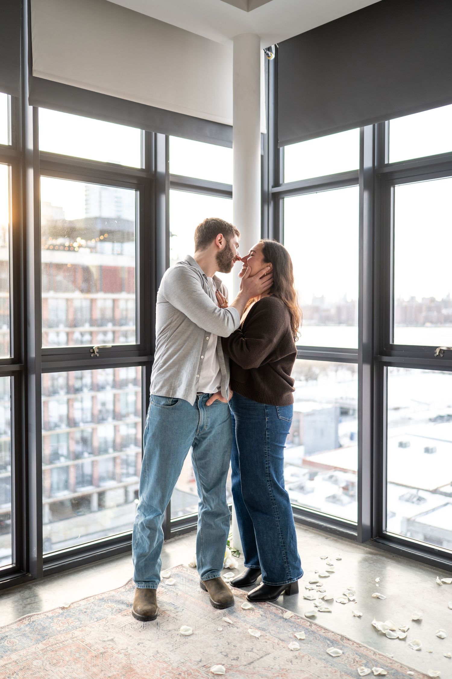 A couple shares an intimate moment by floor-to-ceiling windows overlooking a city view.