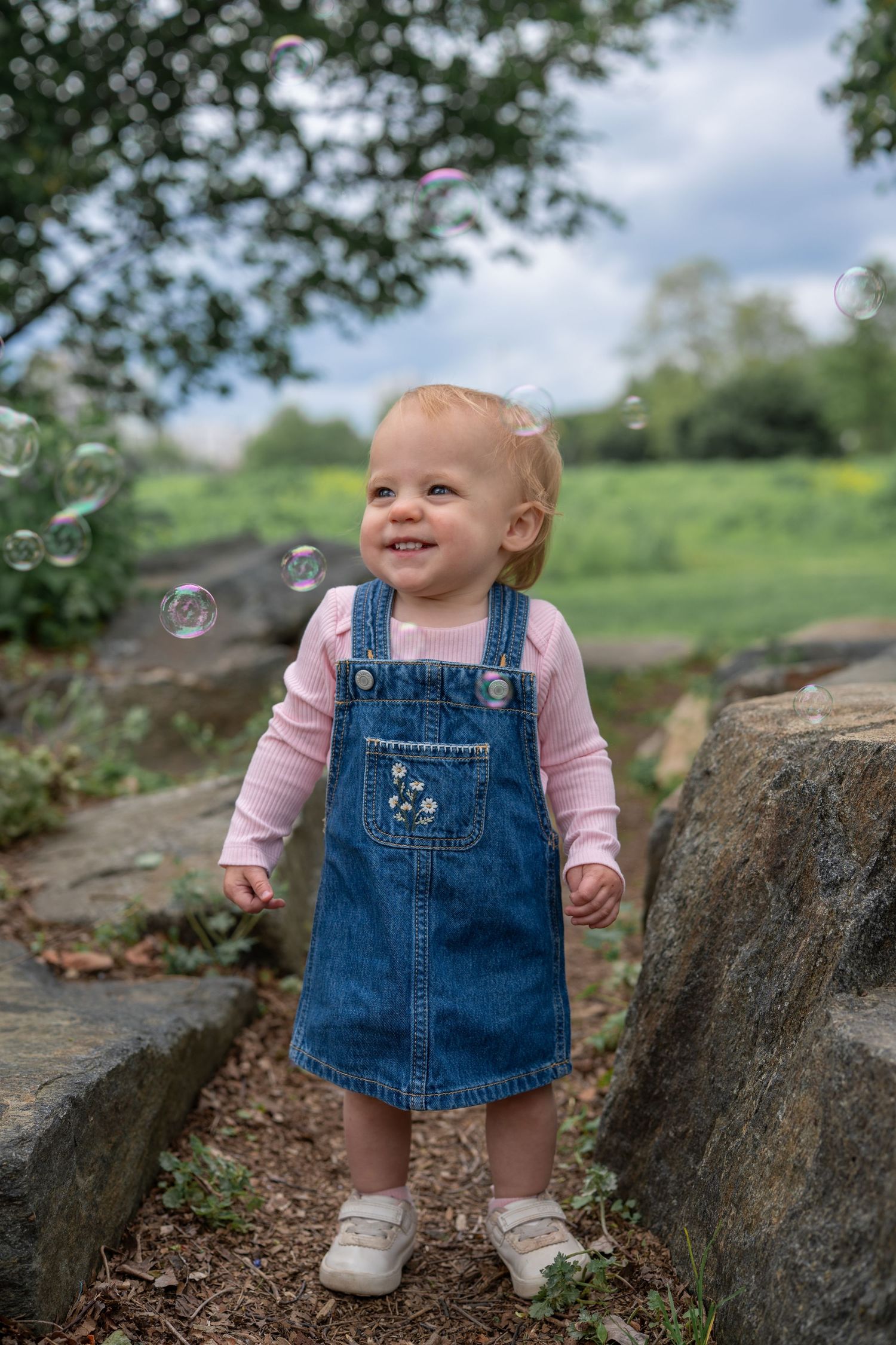 Small child in denim overalls and pink shirt playing with bubbles on rocky terrain.