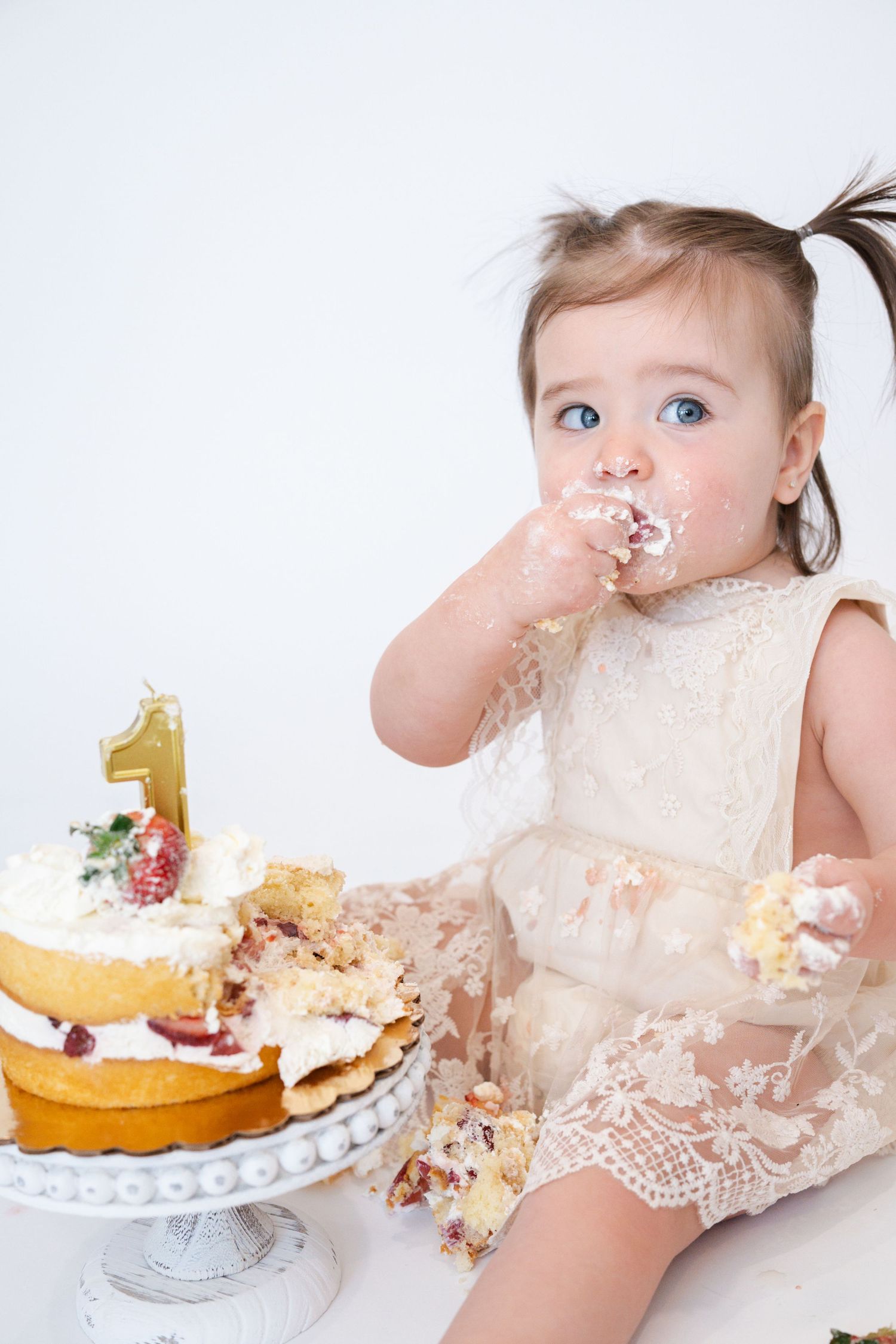 Baby in lace dress enjoying first birthday cake smash with decorated cake on white stand.