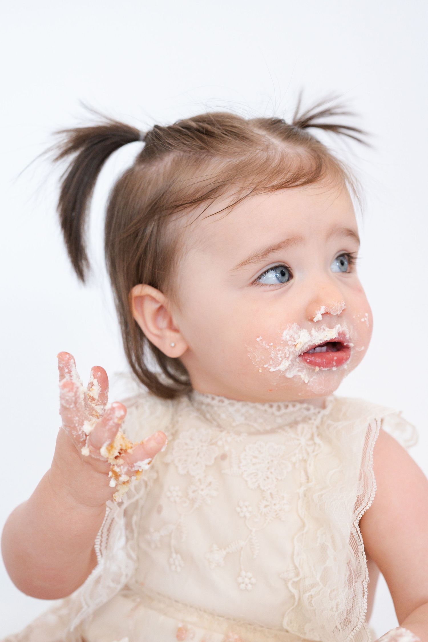 Toddler in lace dress with pigtails has frosting on face and hands while smiling.