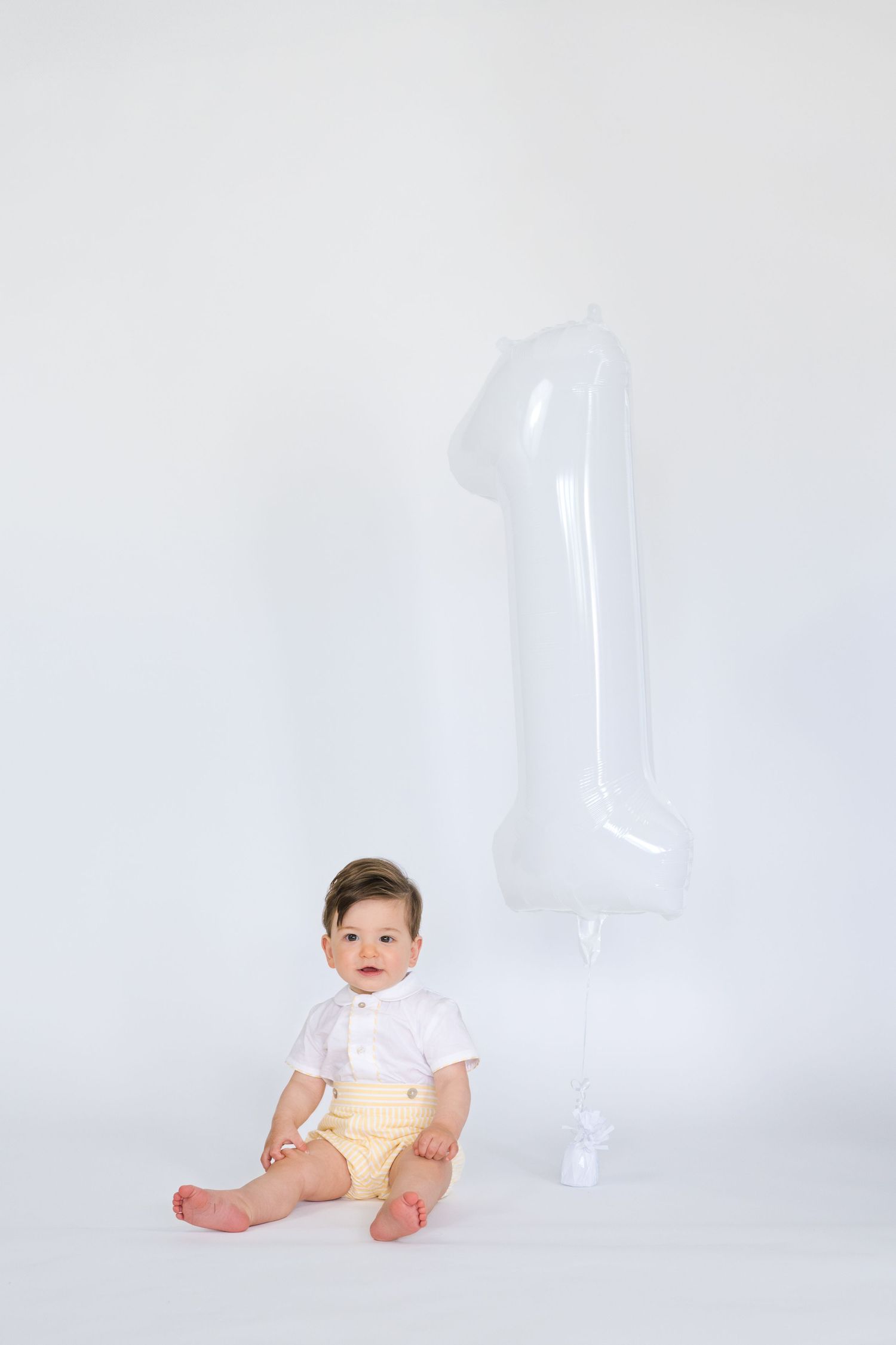 Baby in white and yellow outfit sitting next to transparent number one decoration.