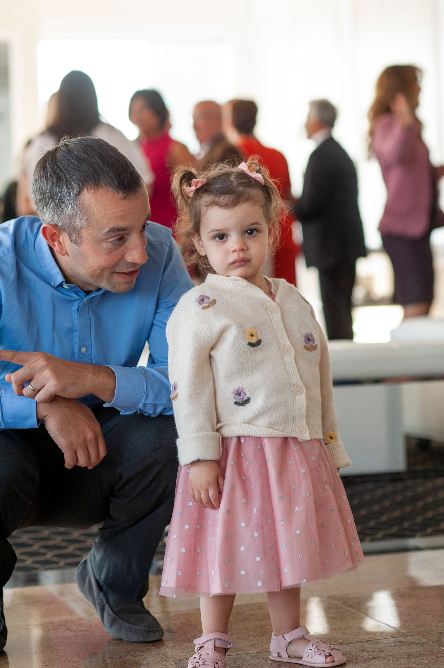 A young child in a pink polka dot dress and white cardigan interacts with an adult in a casual setting.