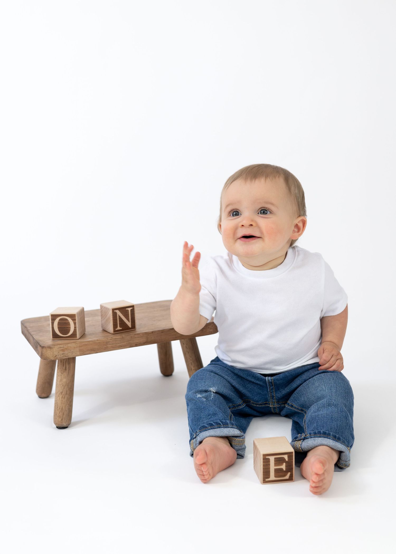 Smiling one-year-old baby boy sitting with wooden letter blocks spelling ONE on white background.