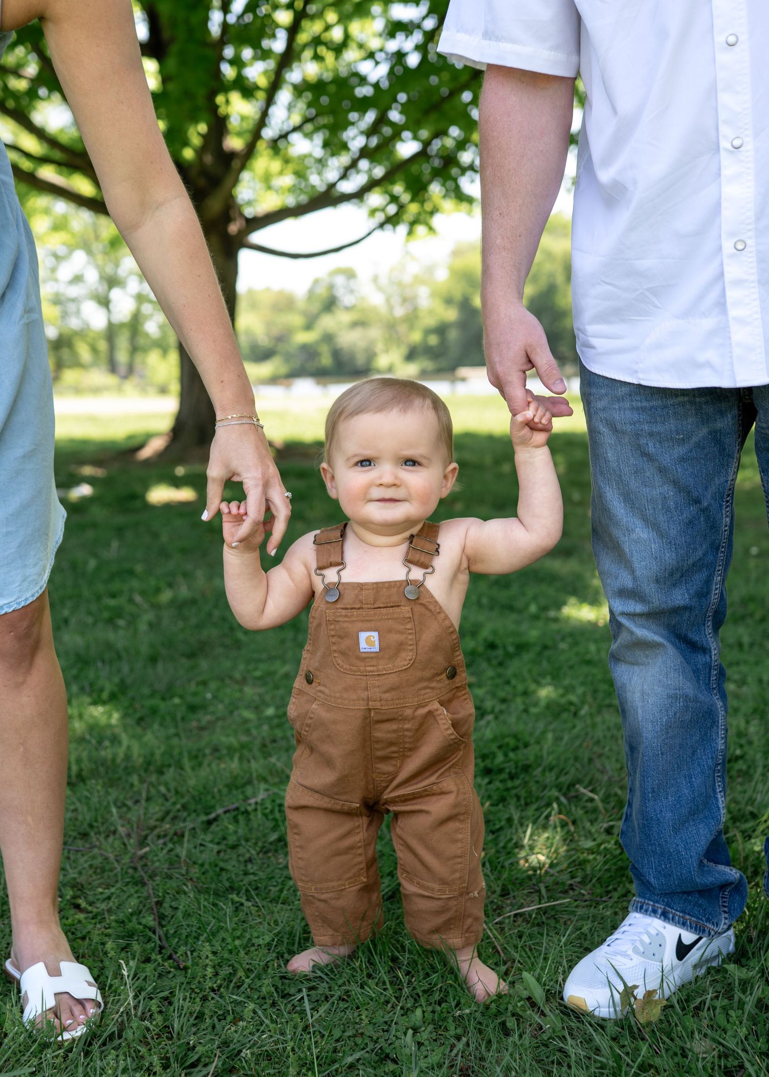 Adorable baby in brown overalls takes first steps while parents hold hands outdoors in a lush park.