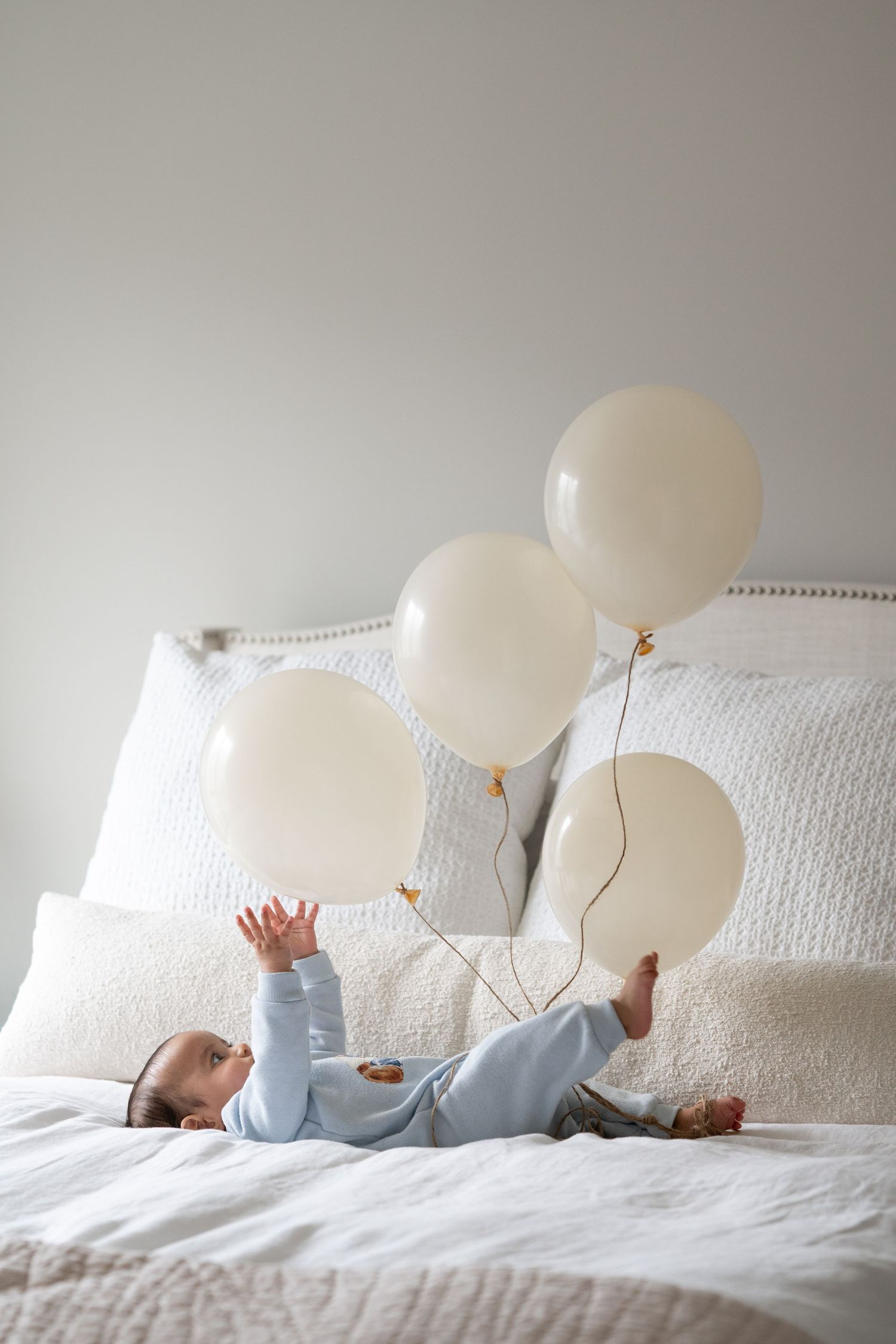 Baby in blue onesie lying on white bed reaching for cream balloons in a bright, minimalist bedroom setting.