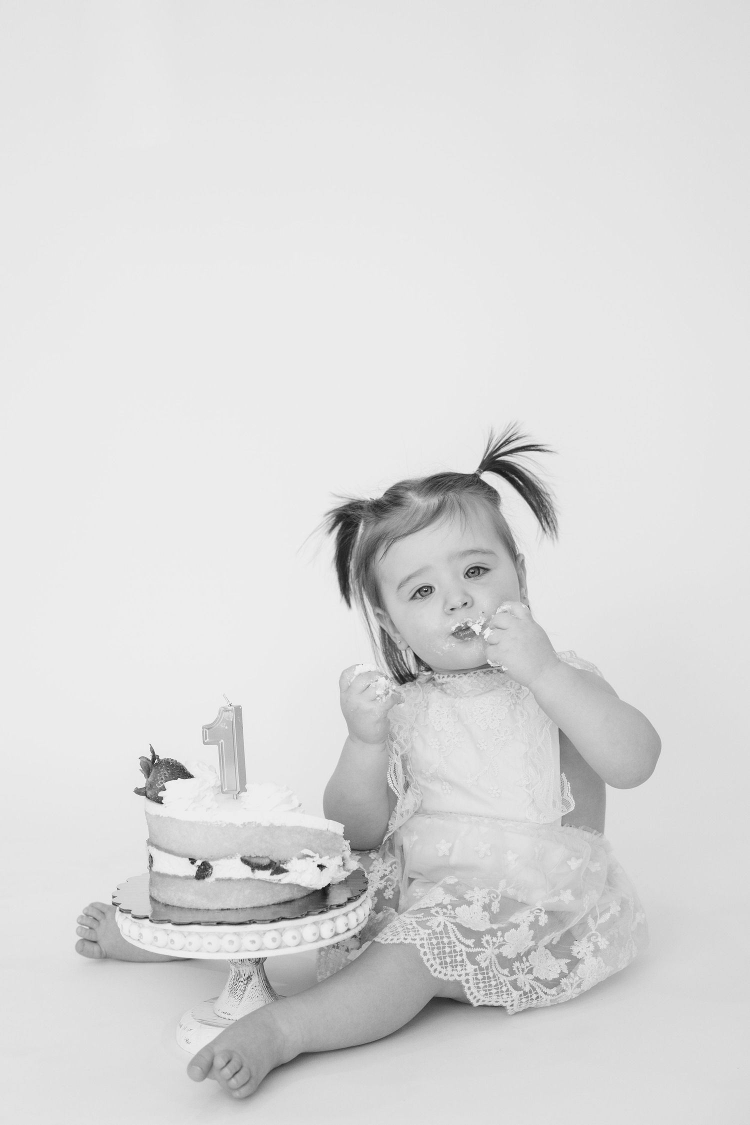 Black and white photo of baby in white dress with first birthday cake.