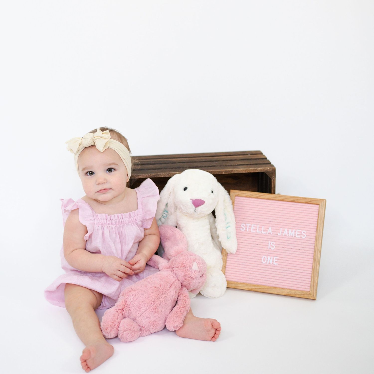 Baby in pink dress sitting with stuffed bunny next to wooden chest and letter board.