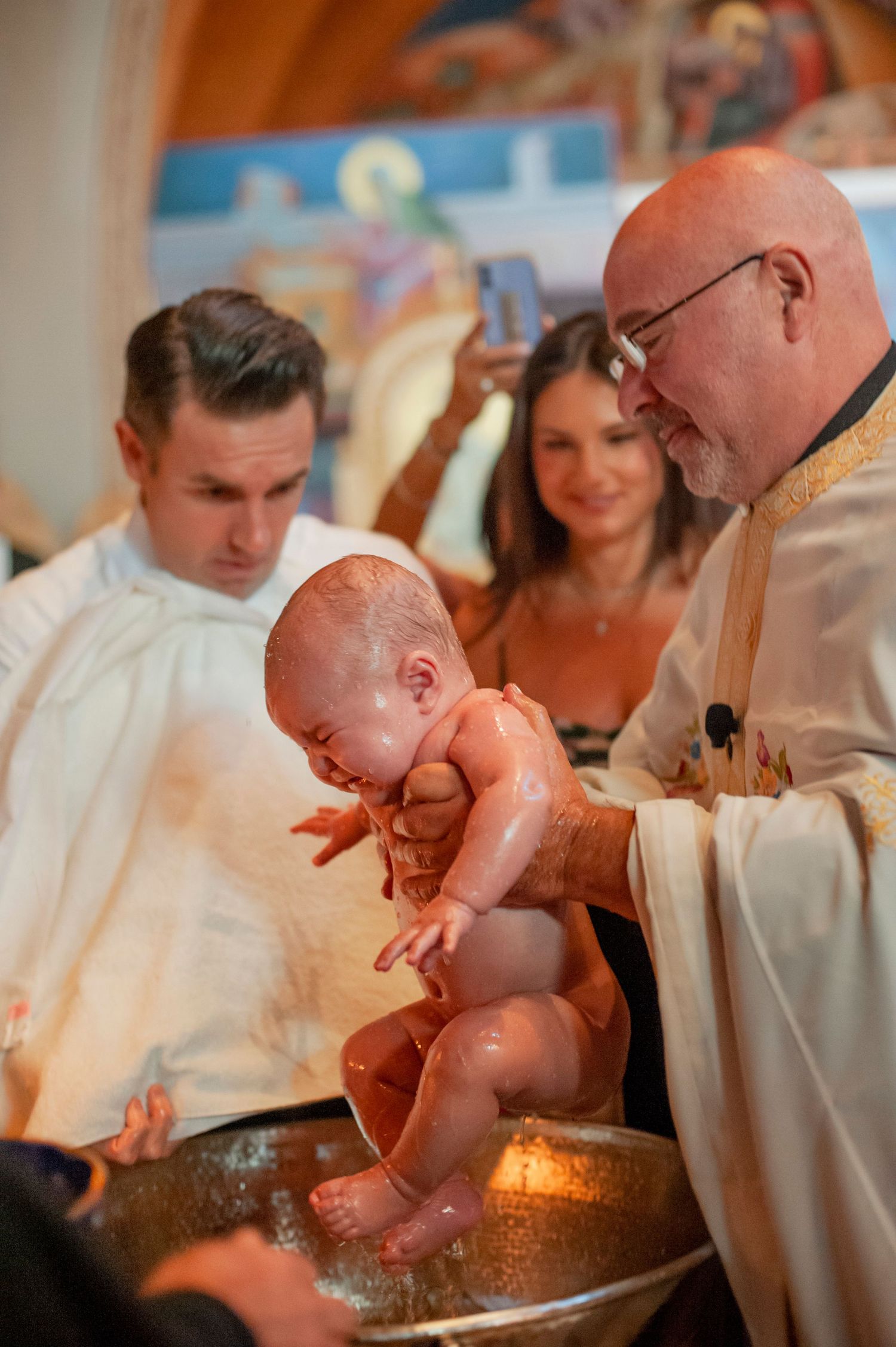 A baptism ceremony is performed with water at a church font while family members look on.