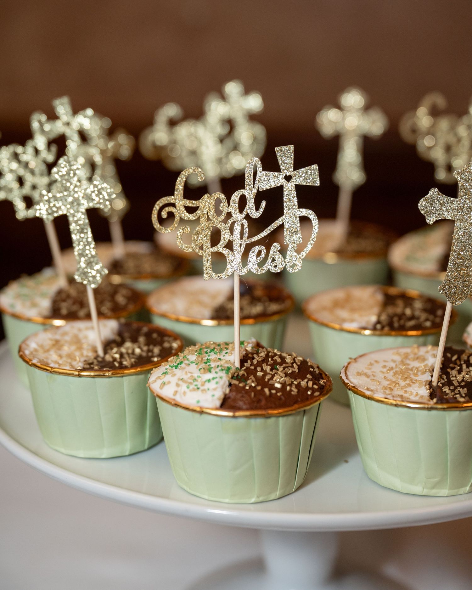 Cupcakes decorated with silver cross toppers and 'God Bless' text arranged on a white cake stand.