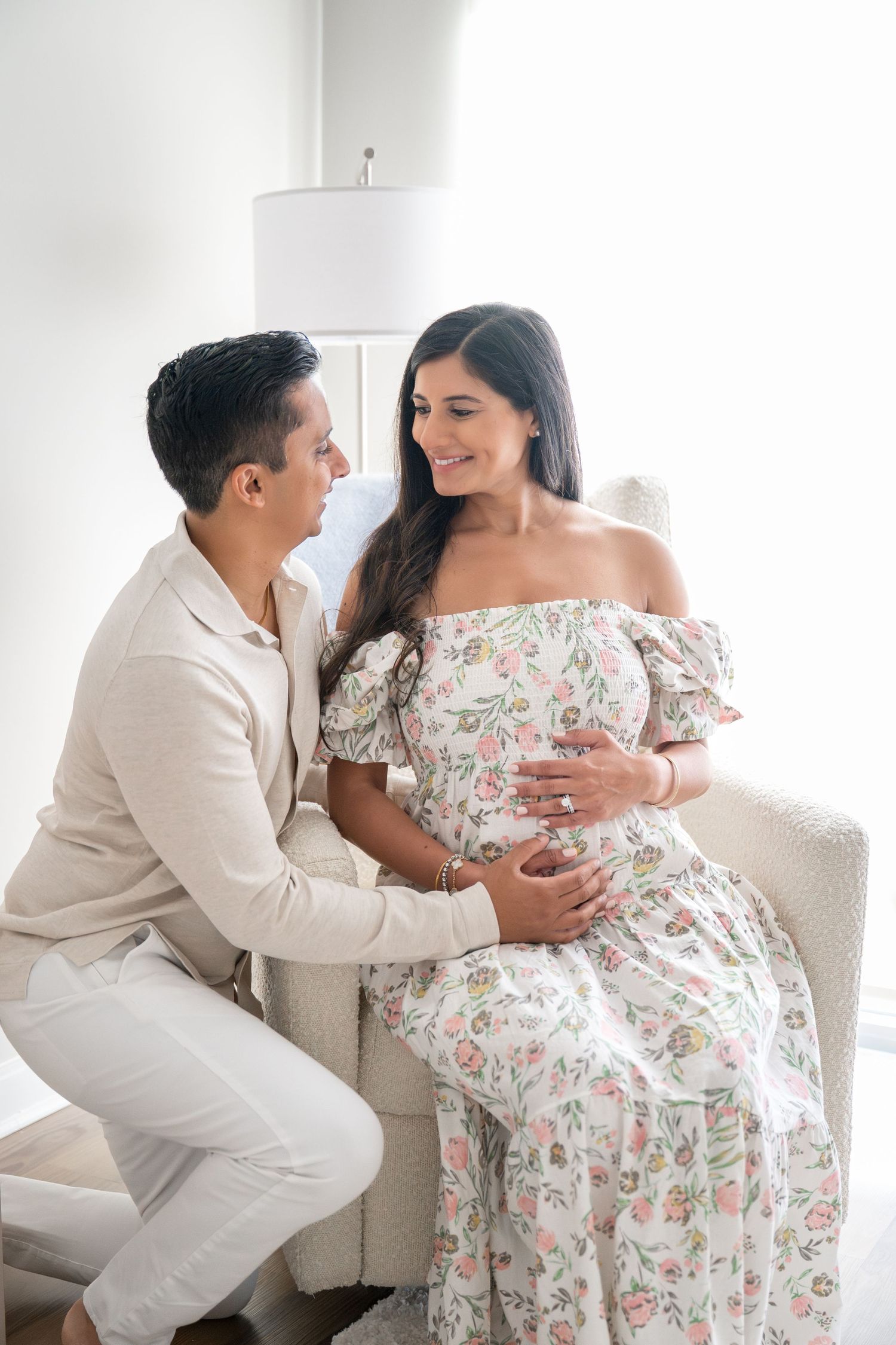 A couple shares an intimate moment in a bright room, with her wearing a floral off-shoulder dress.