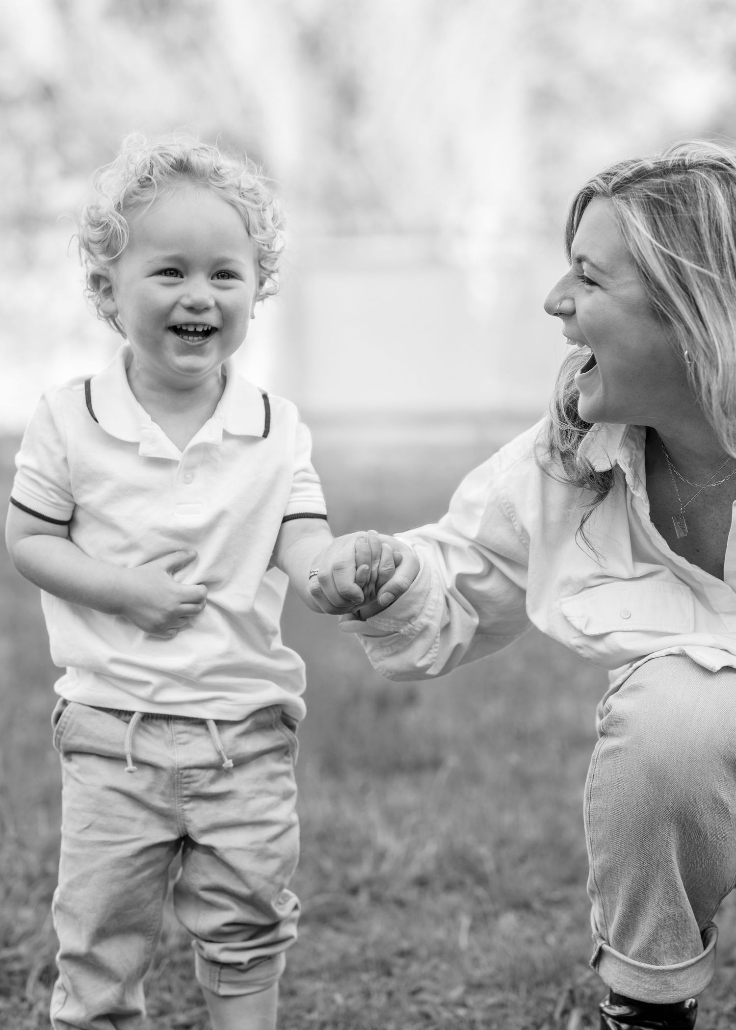 Black and white photo of a laughing toddler boy holding hands with a smiling woman outdoors.