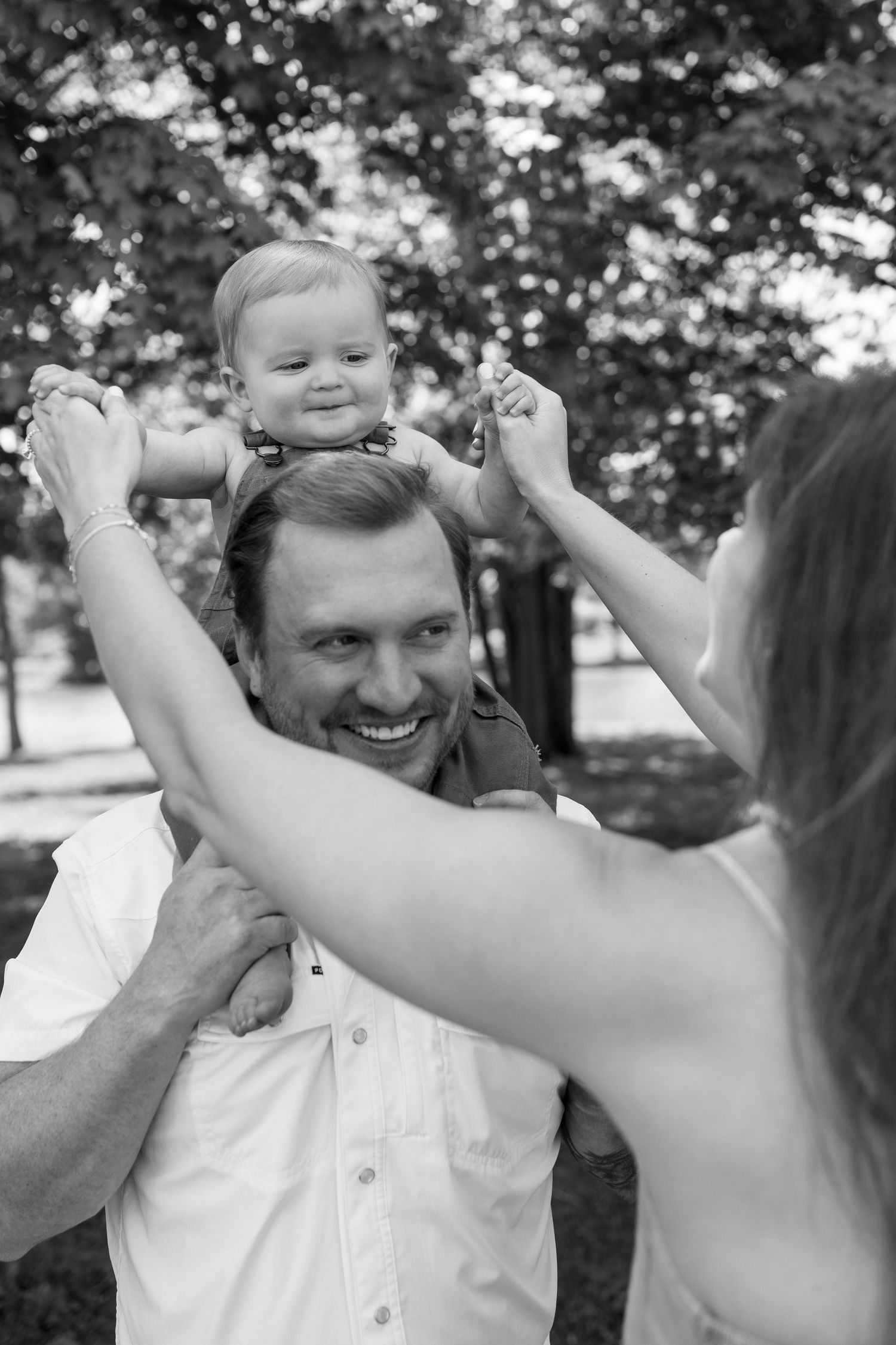 Black and white photo of smiling dad holding baby on shoulders while mom assists outdoors in park.
