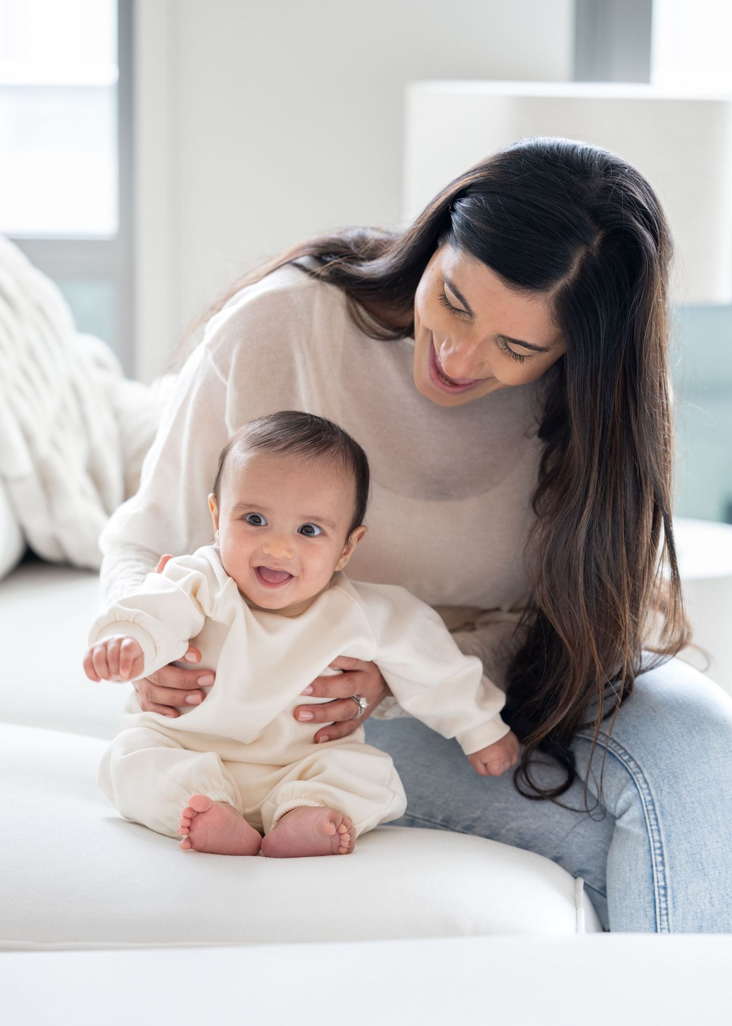 Smiling mother in white sweater helps her happy baby sit up on a white couch indoors.