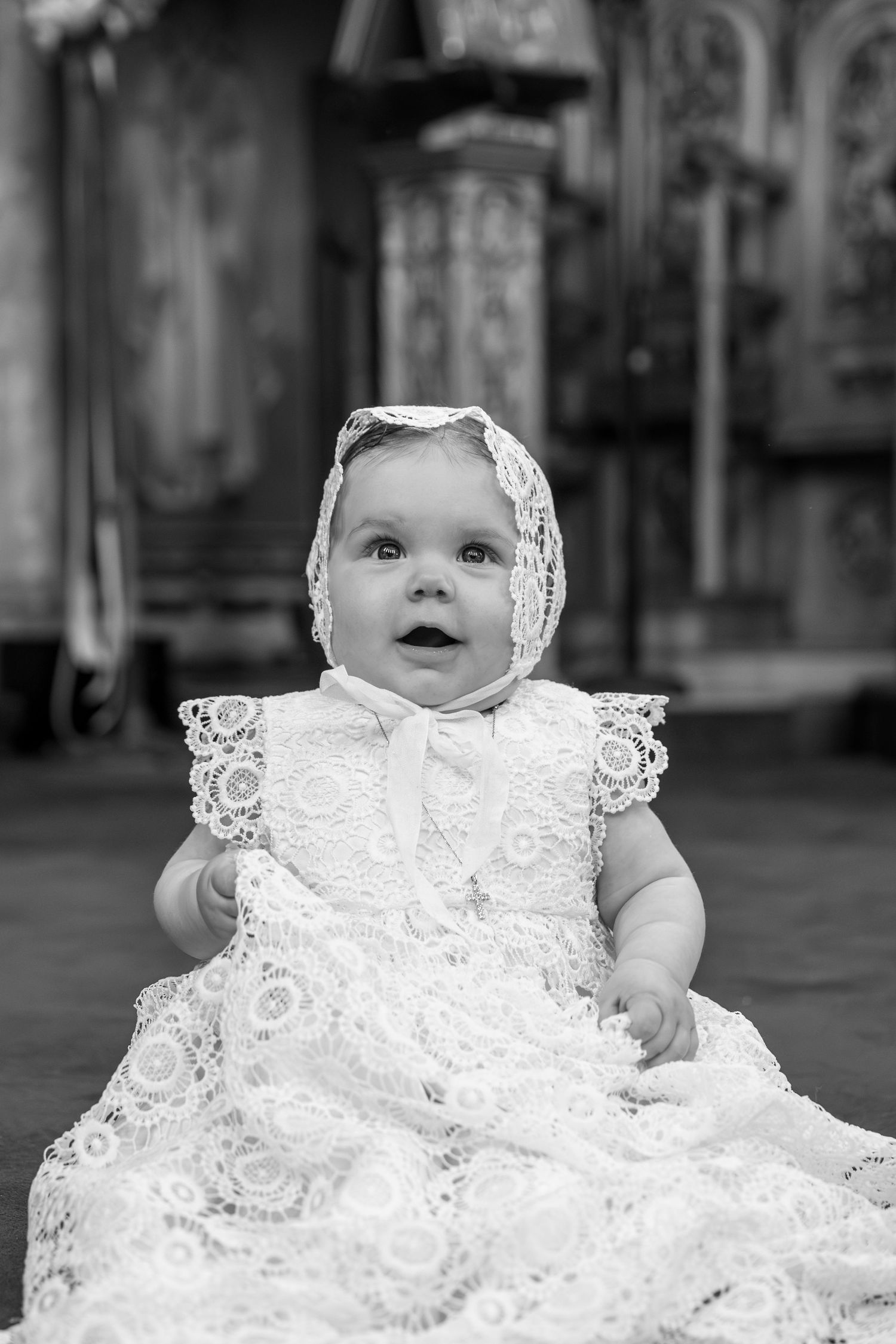 Baby in white christening gown and bonnet sitting during baptism ceremony in church.