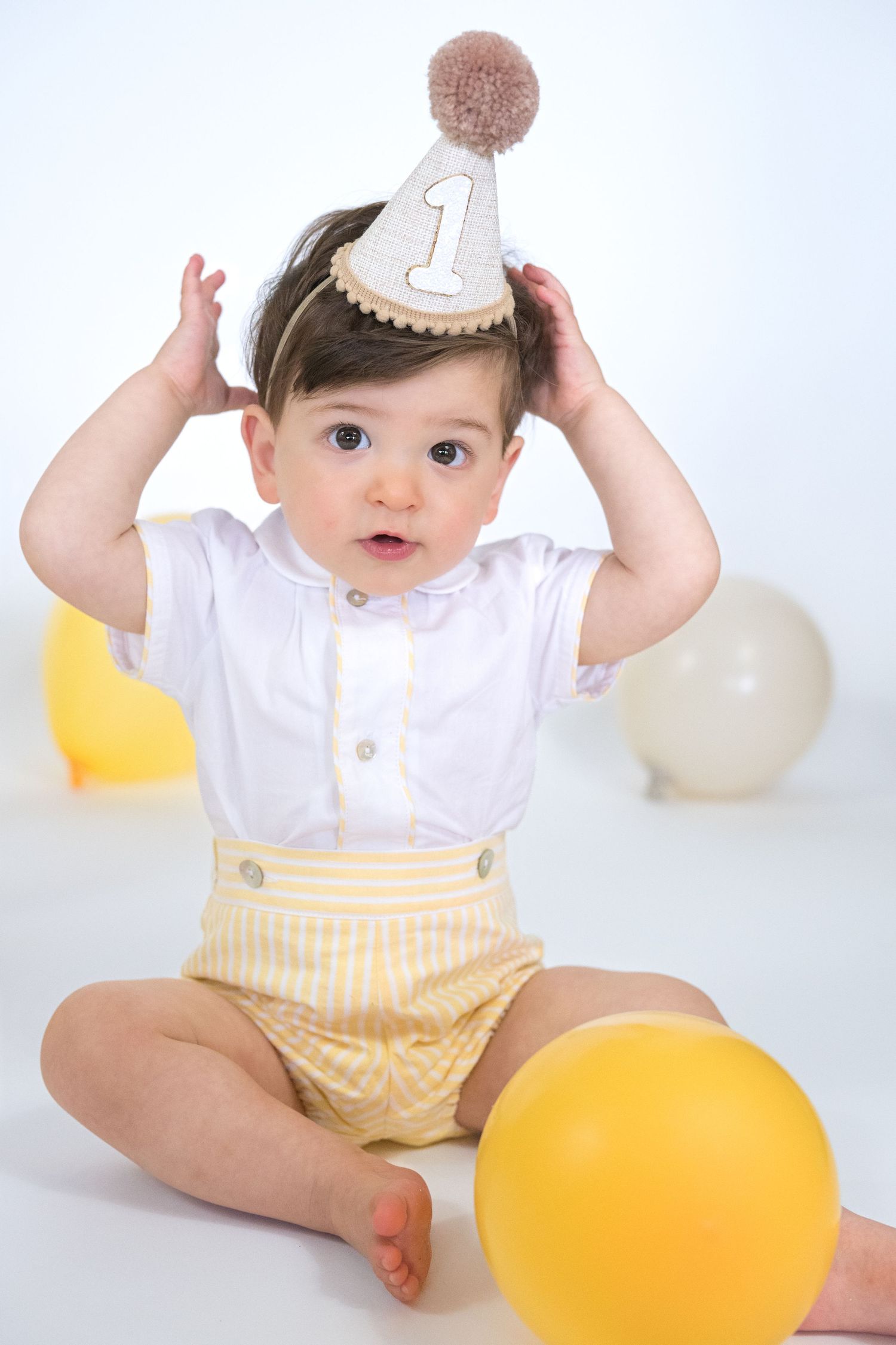 Baby in white shirt and yellow shorts sits with balloons and party decorations for first birthday celebration.
