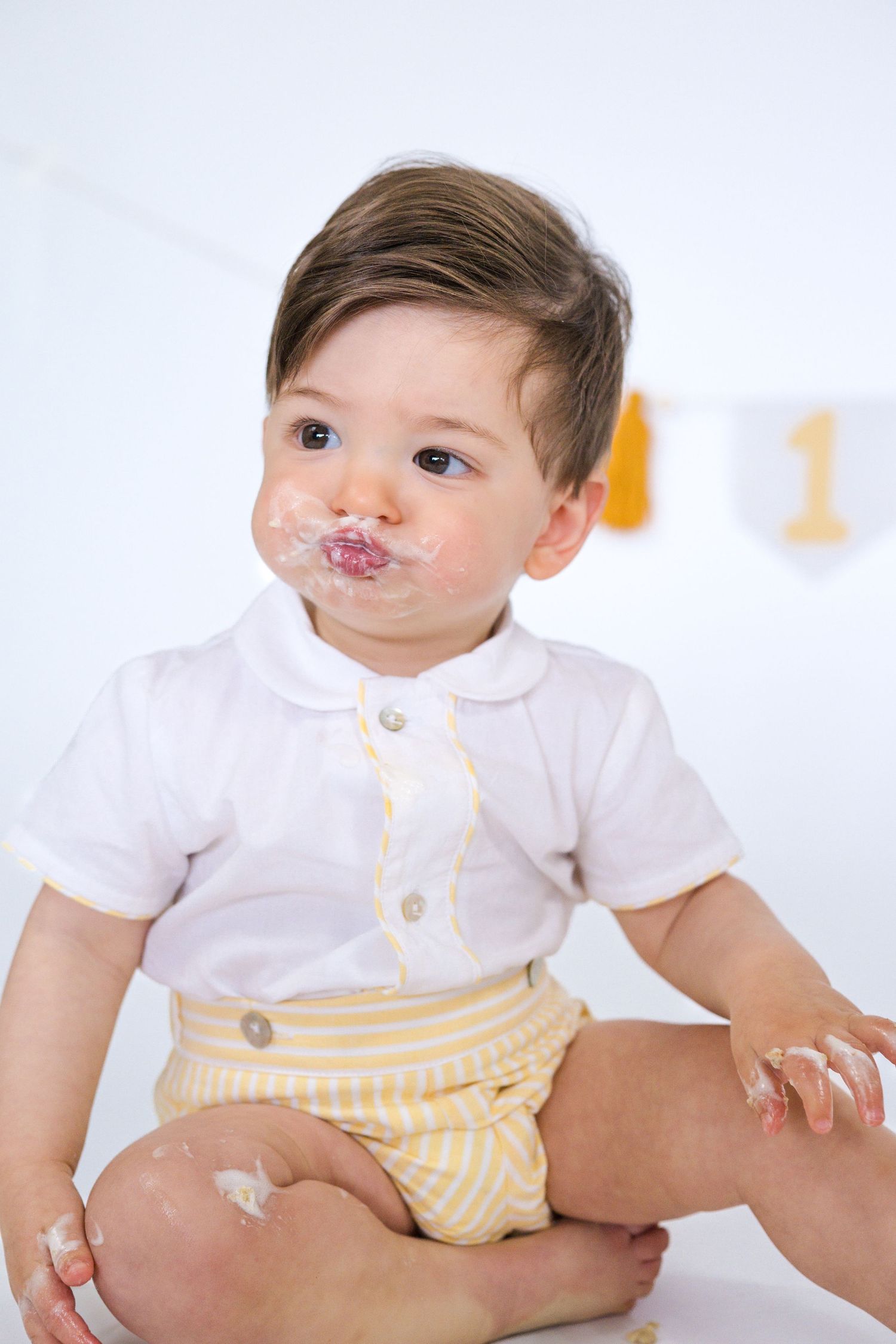 Baby in white collared shirt and yellow shorts smiles with cake-covered face.