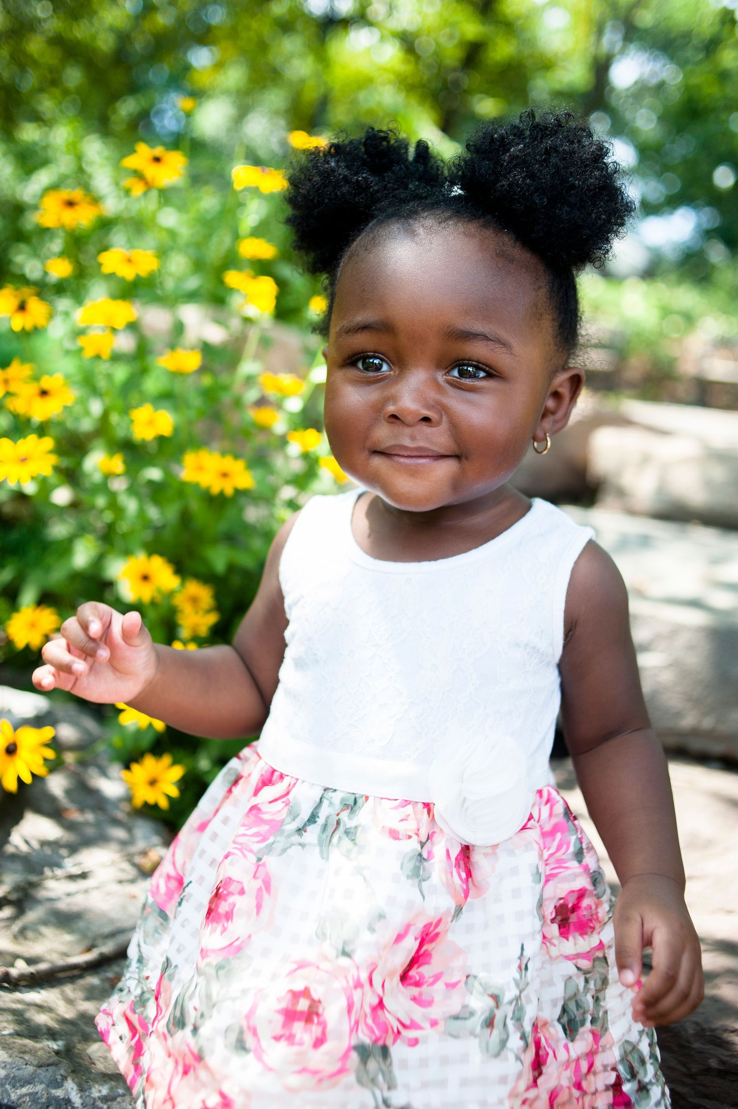 Toddler in a white and floral dress poses near yellow flowers in a garden.