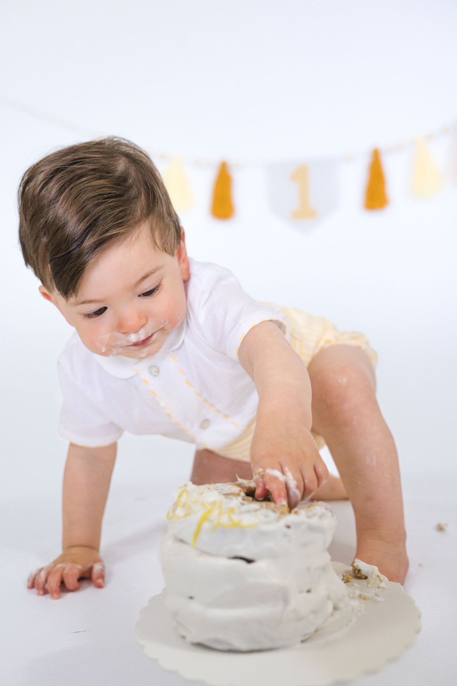 Baby in white shirt smashes and plays with birthday cake during cake smash photo session with yellow banner decor.