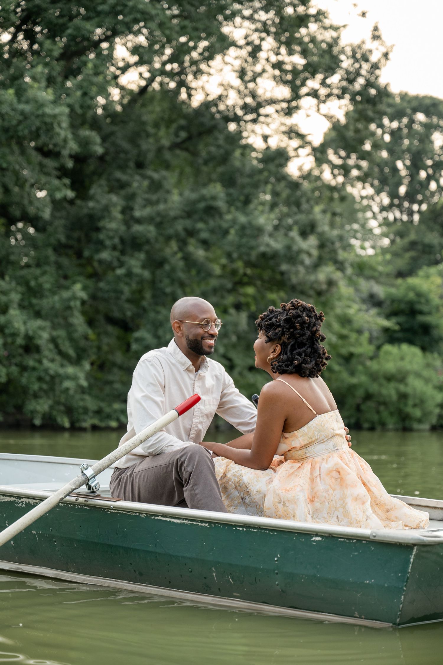 A couple shares a romantic moment while sitting in a green rowboat on a tranquil lake during sunset.