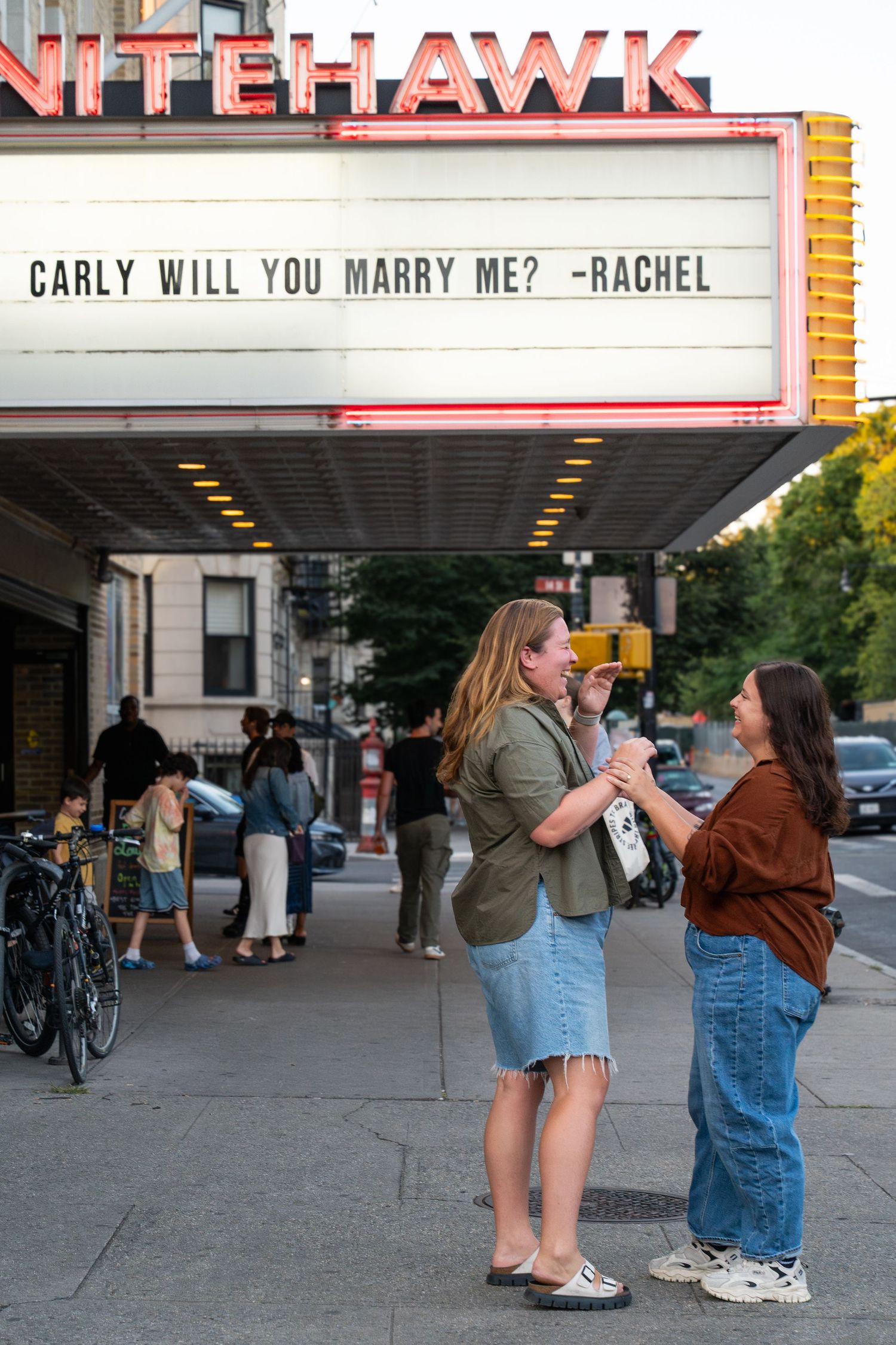 Proposal scene outside Ritehawk theater with marquee sign reading 'CARLY WILL YOU MARRY ME? -RACHEL' at dusk.