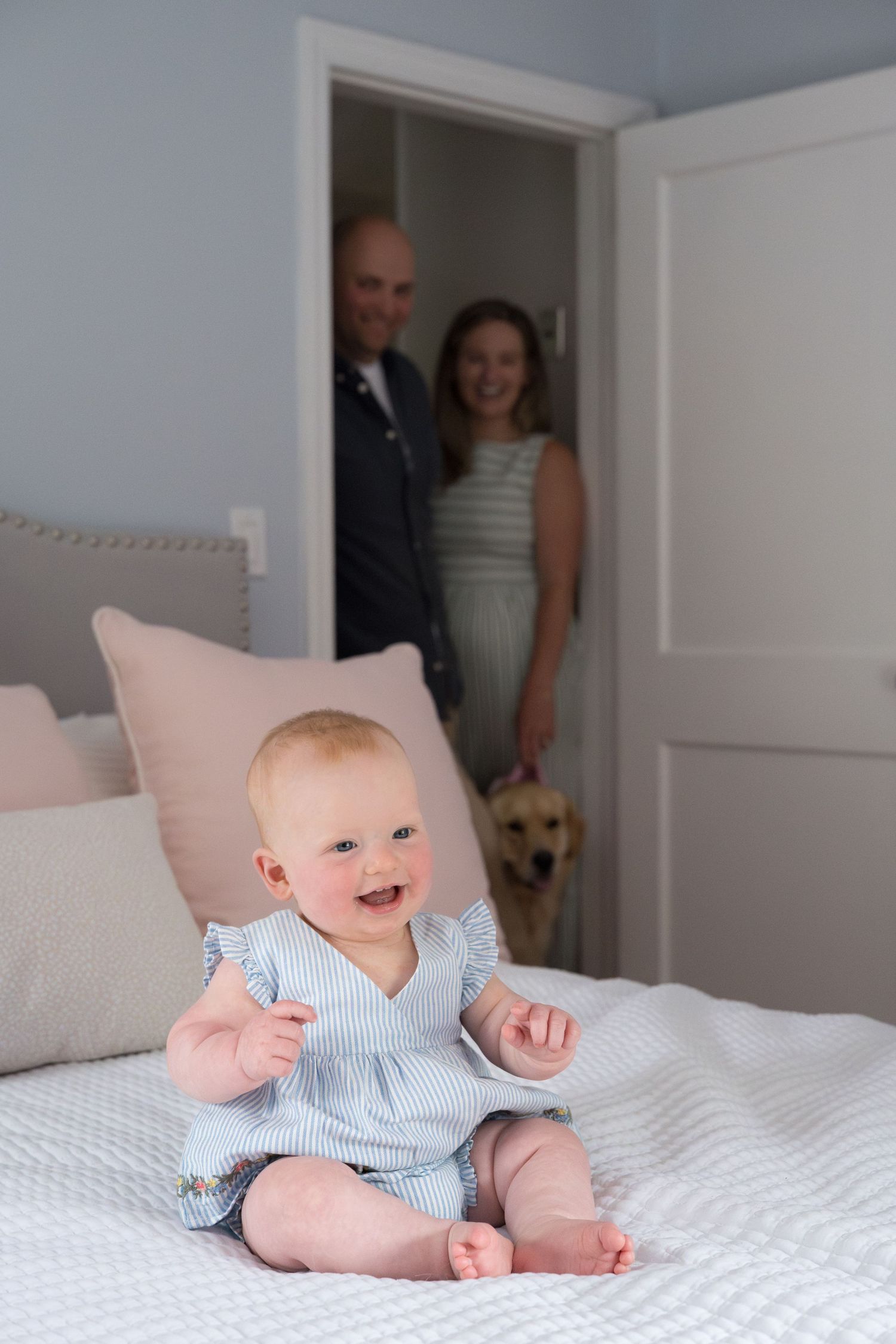 A happy baby in blue striped romper sits on a white bed while family watches from doorway.
