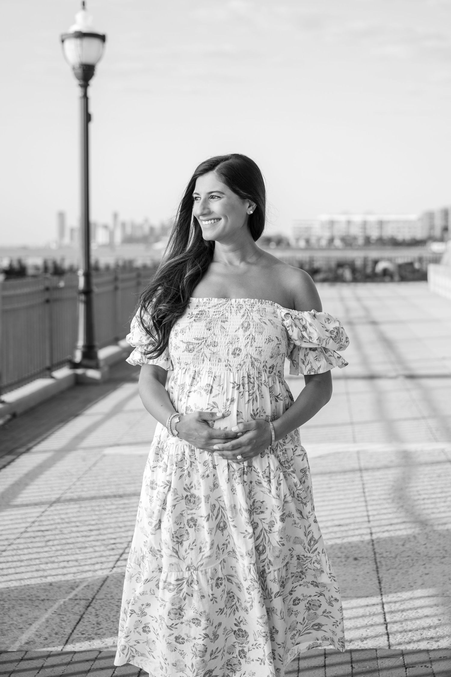 A woman in a floral dress twirls joyfully on a waterfront promenade with city views.