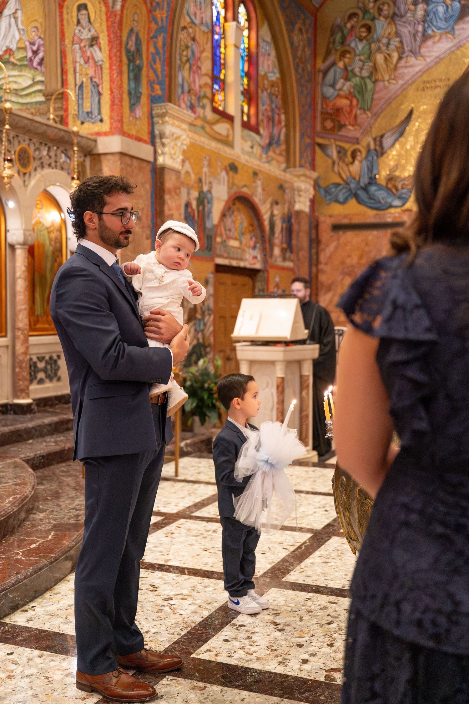 A religious baptism ceremony unfolds in a beautifully decorated church with Byzantine-style murals on the walls.