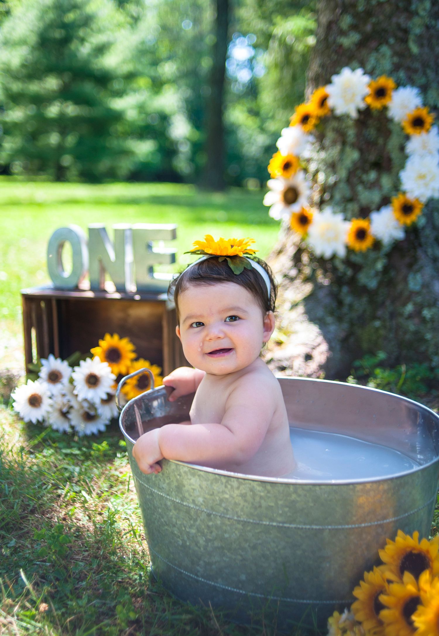 Baby taking milk bath in metal tub surrounded by sunflowers and ONE sign decoration.
