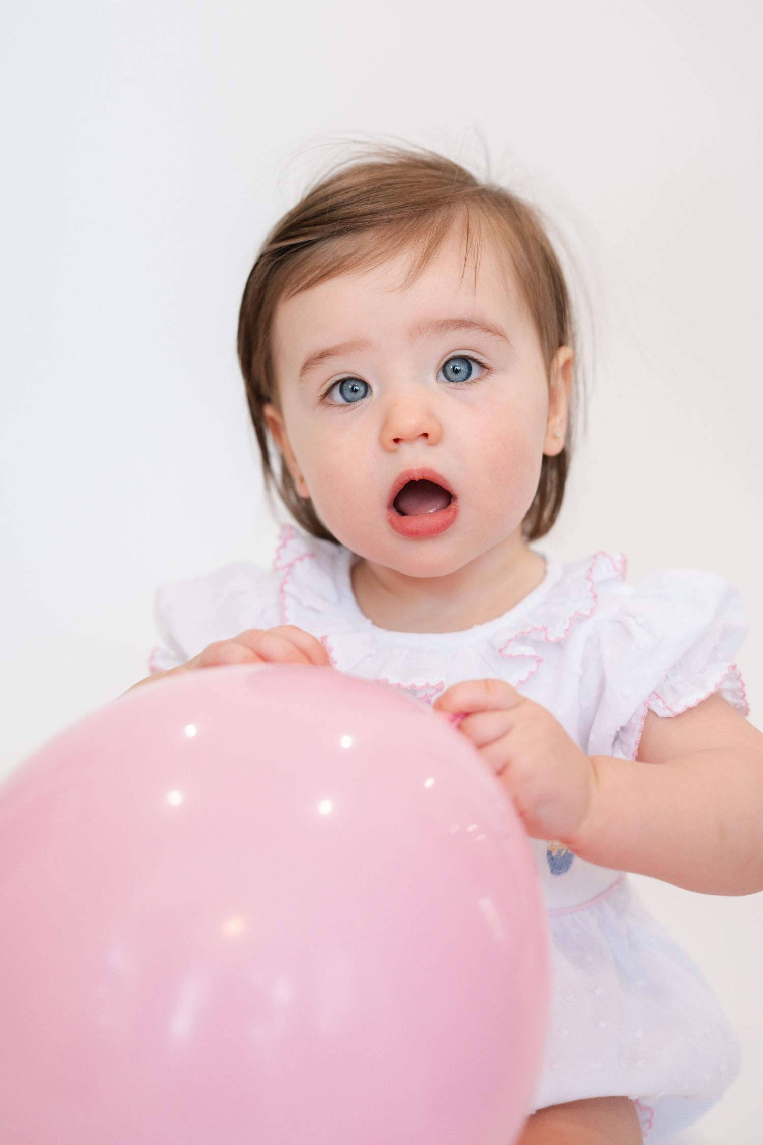 Baby in white dress playing with pink balloon against white backdrop.