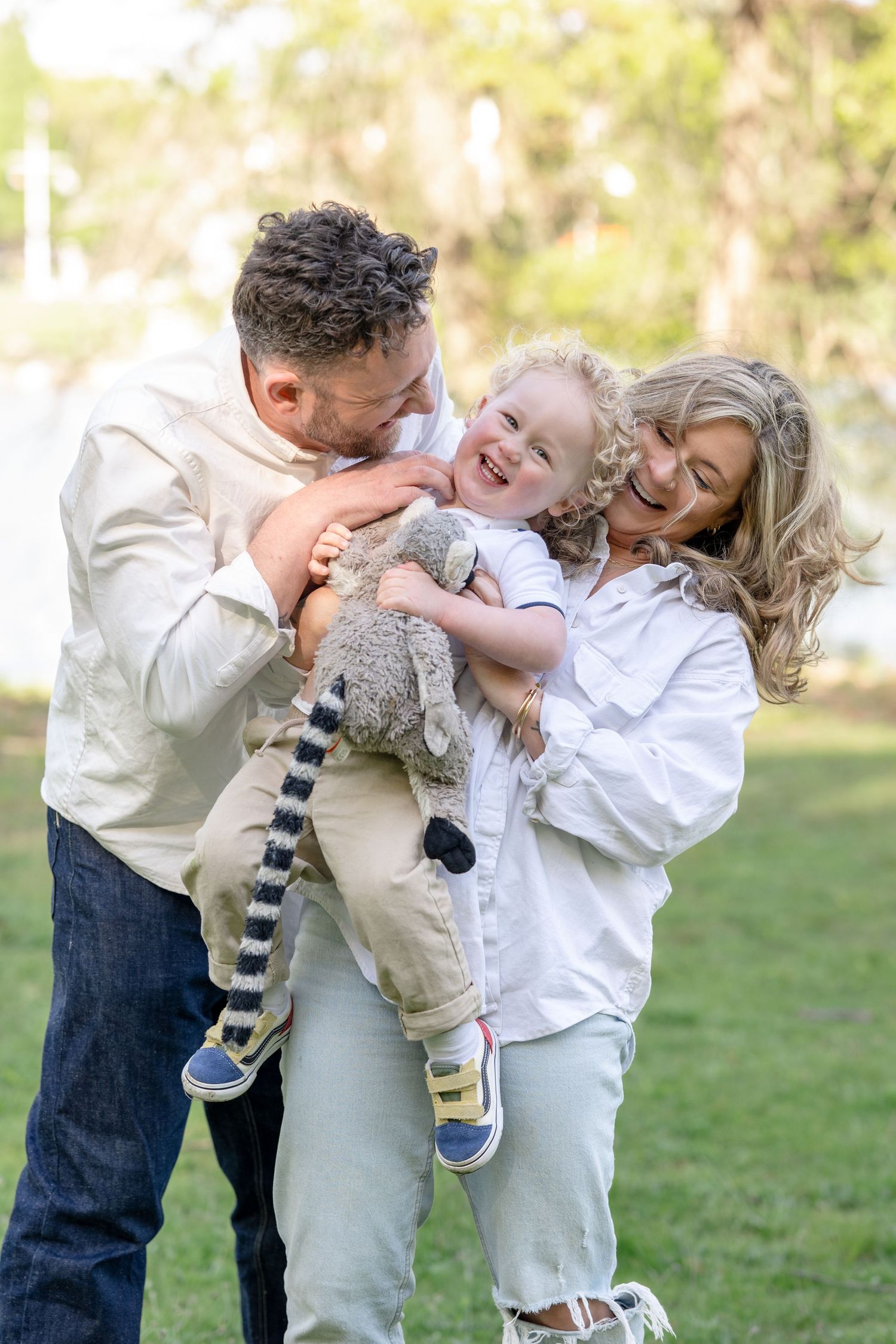 Happy family with parents kissing and hugging laughing toddler holding stuffed animal toy outdoors in green park.