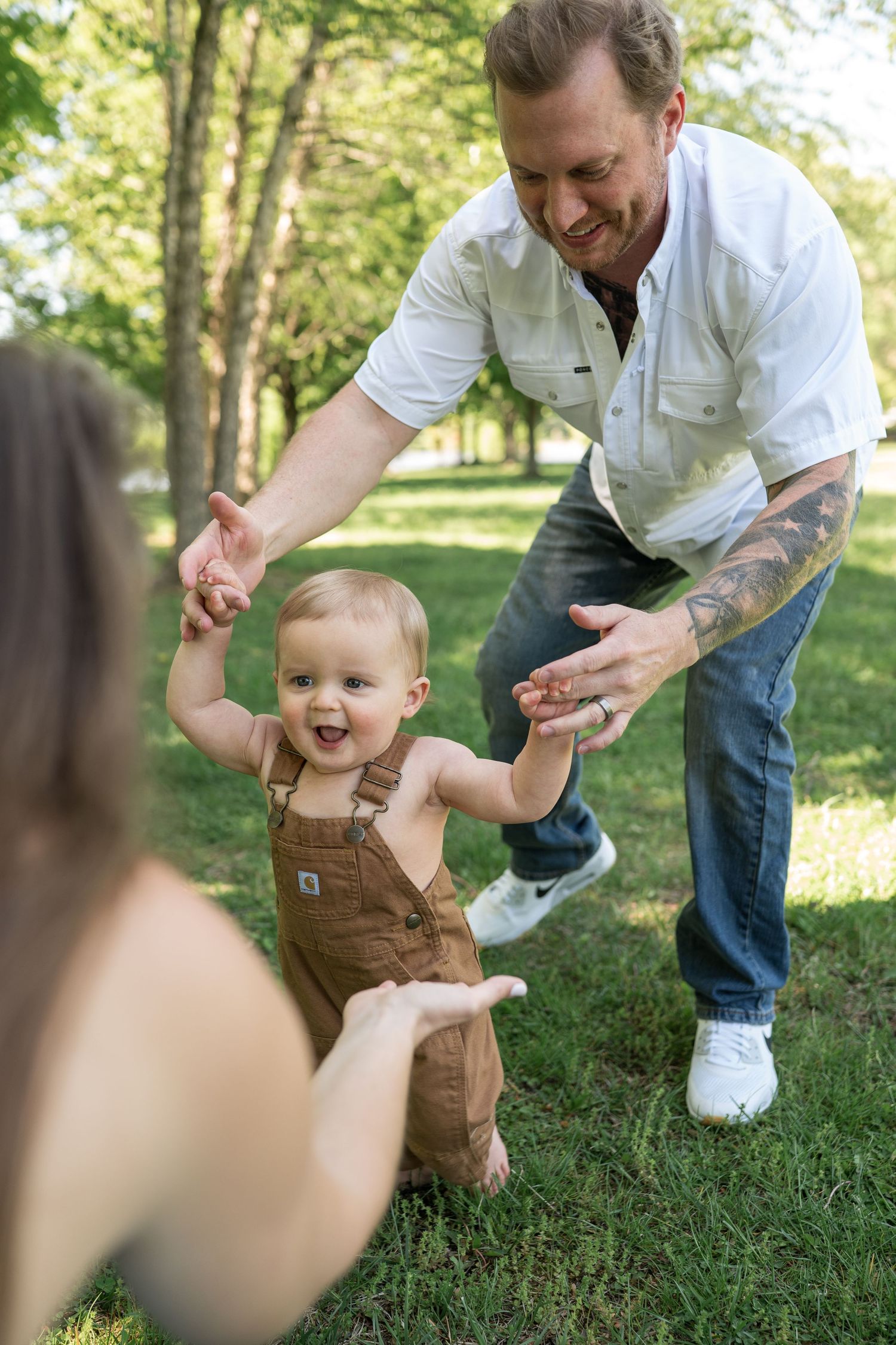 A smiling baby in brown overalls takes first steps outdoors, supported by a tattooed father in a sunny green park.