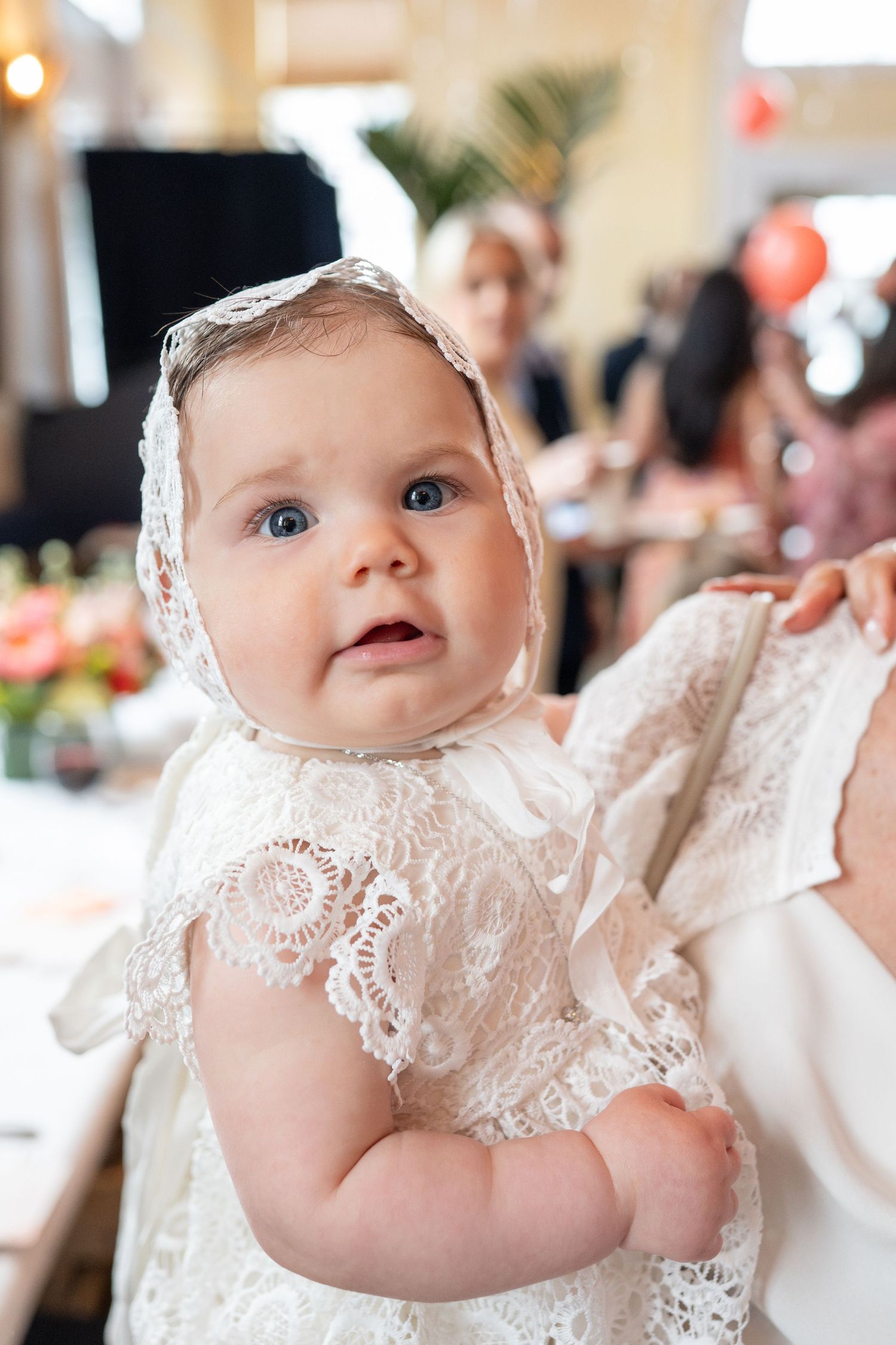 Adorable baby in a white lace christening gown and bonnet with blue eyes at a celebration event.