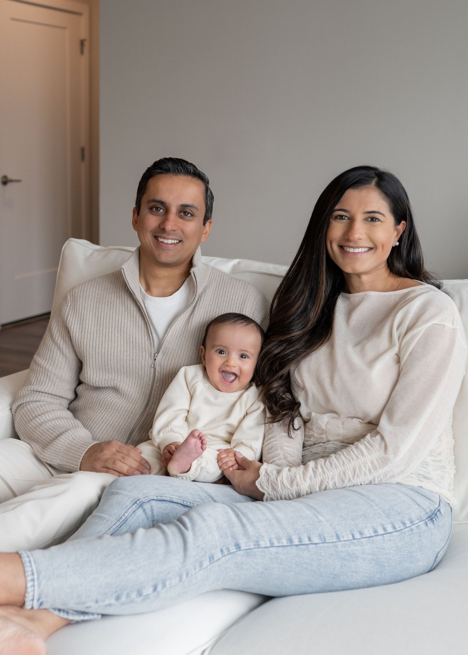 Happy family of three with baby in cream outfit sitting together on white couch at home.