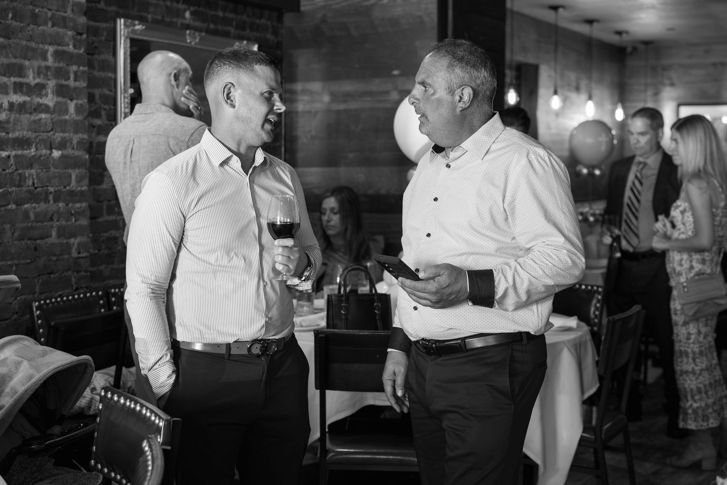 Two people engaged in conversation at a social gathering while holding drinks in a dimly lit room with brick walls.