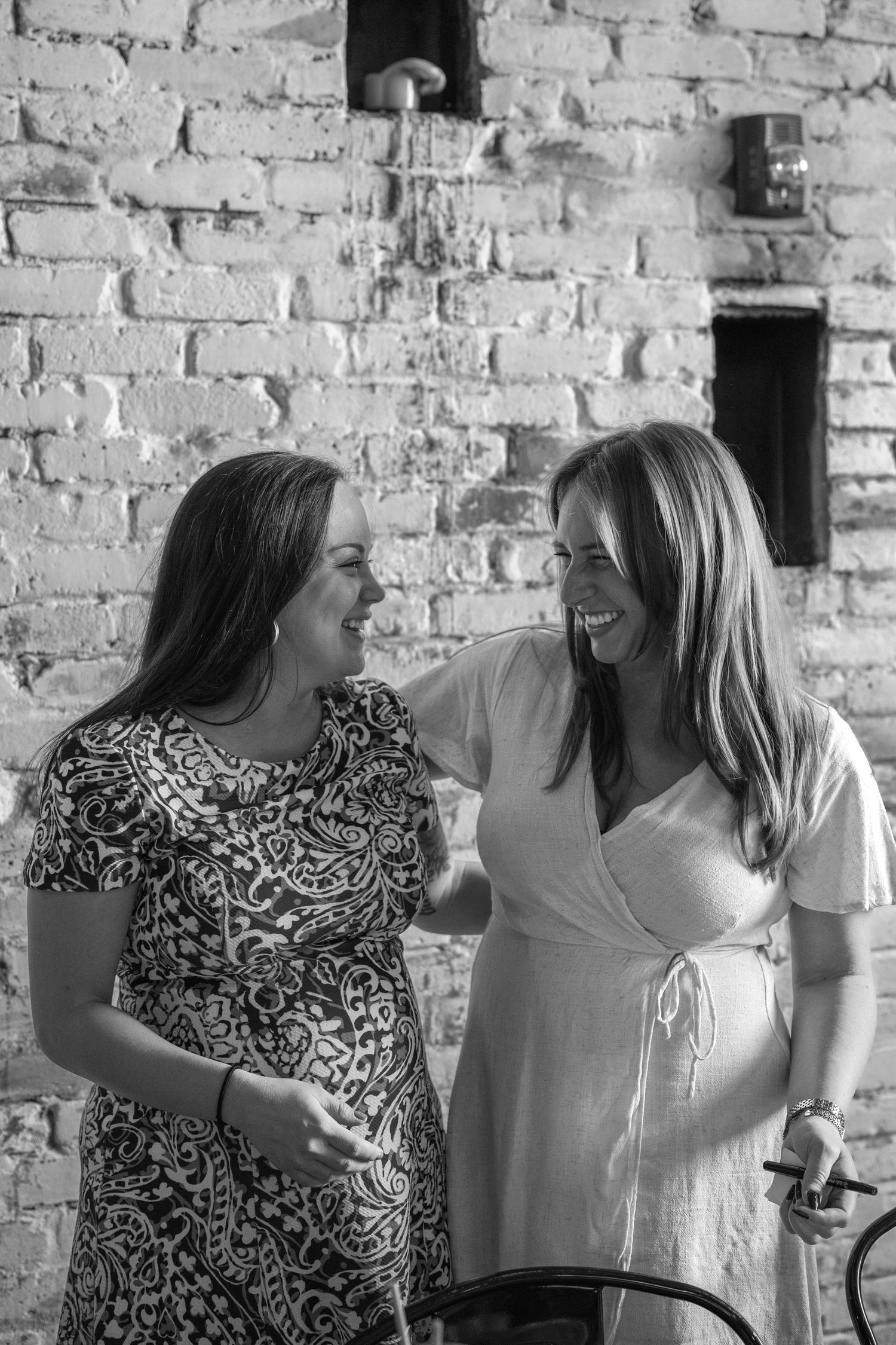 Two women share a candid laughing moment against a rustic brick wall background in black and white.