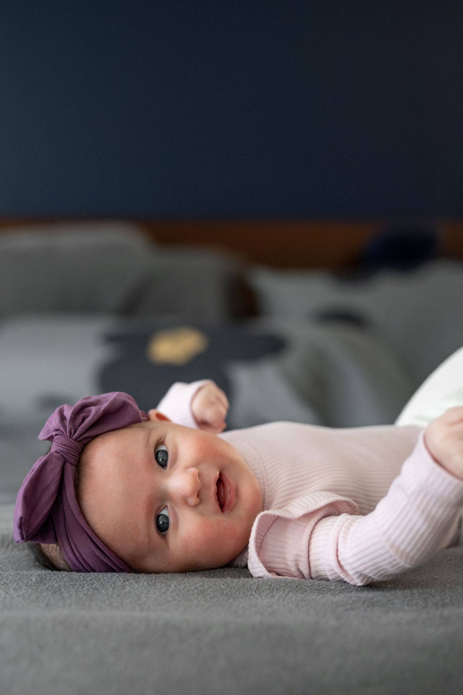 A baby in a white outfit and purple headband lies on a gray surface looking at the camera.