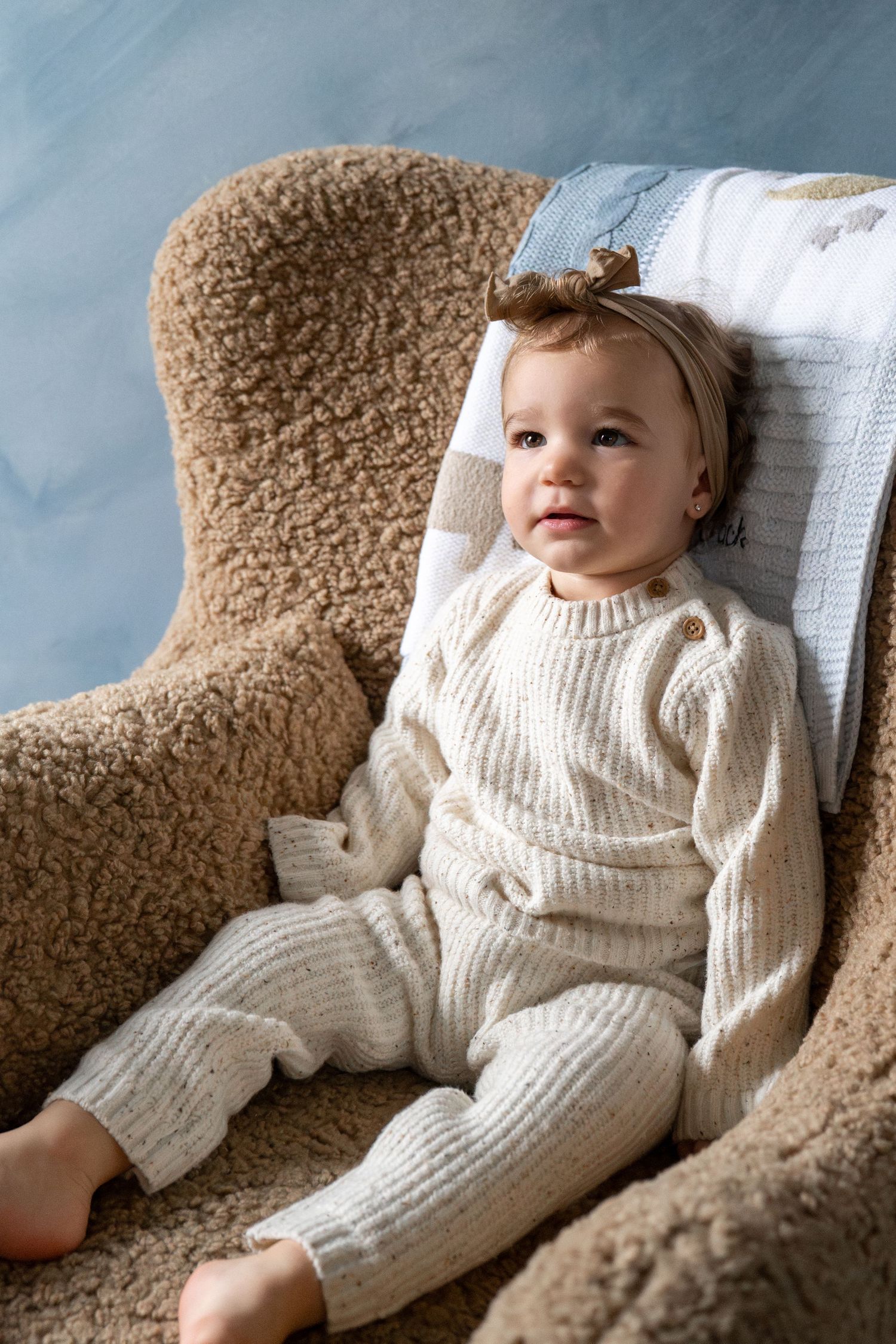 A figure wearing a cozy white knit outfit reclines on a fluffy brown chair near a window.