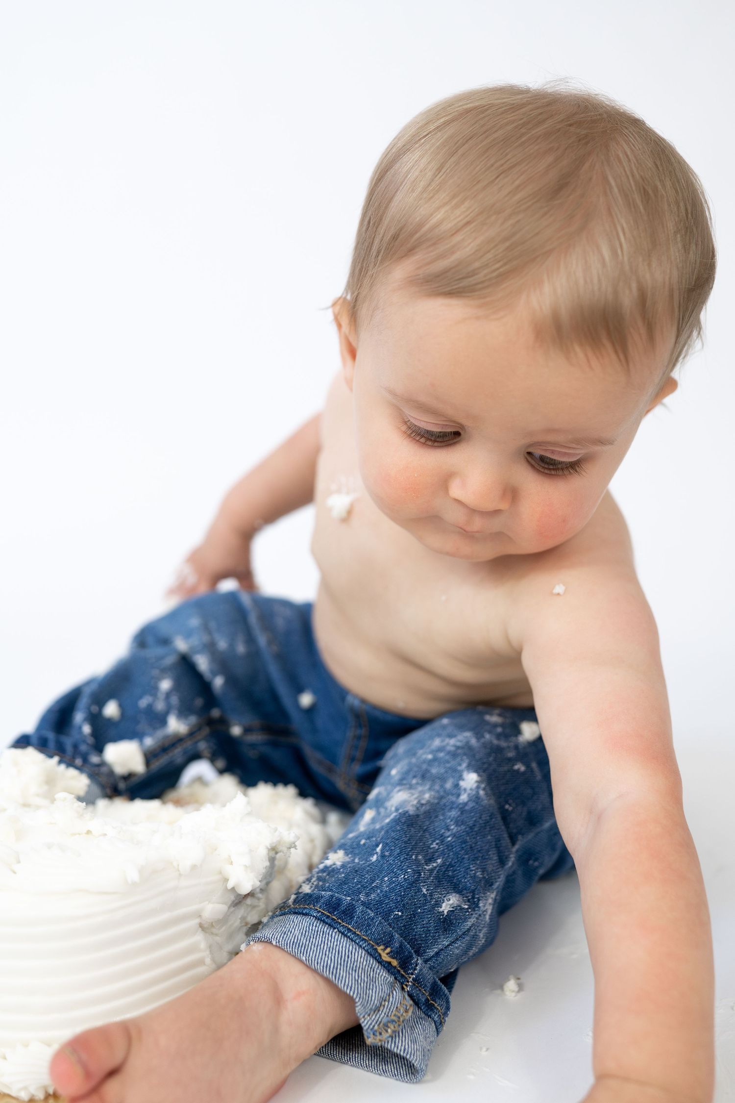 Baby in jeans smashing a white cake, covered in frosting, during a first birthday cake smash photo session.