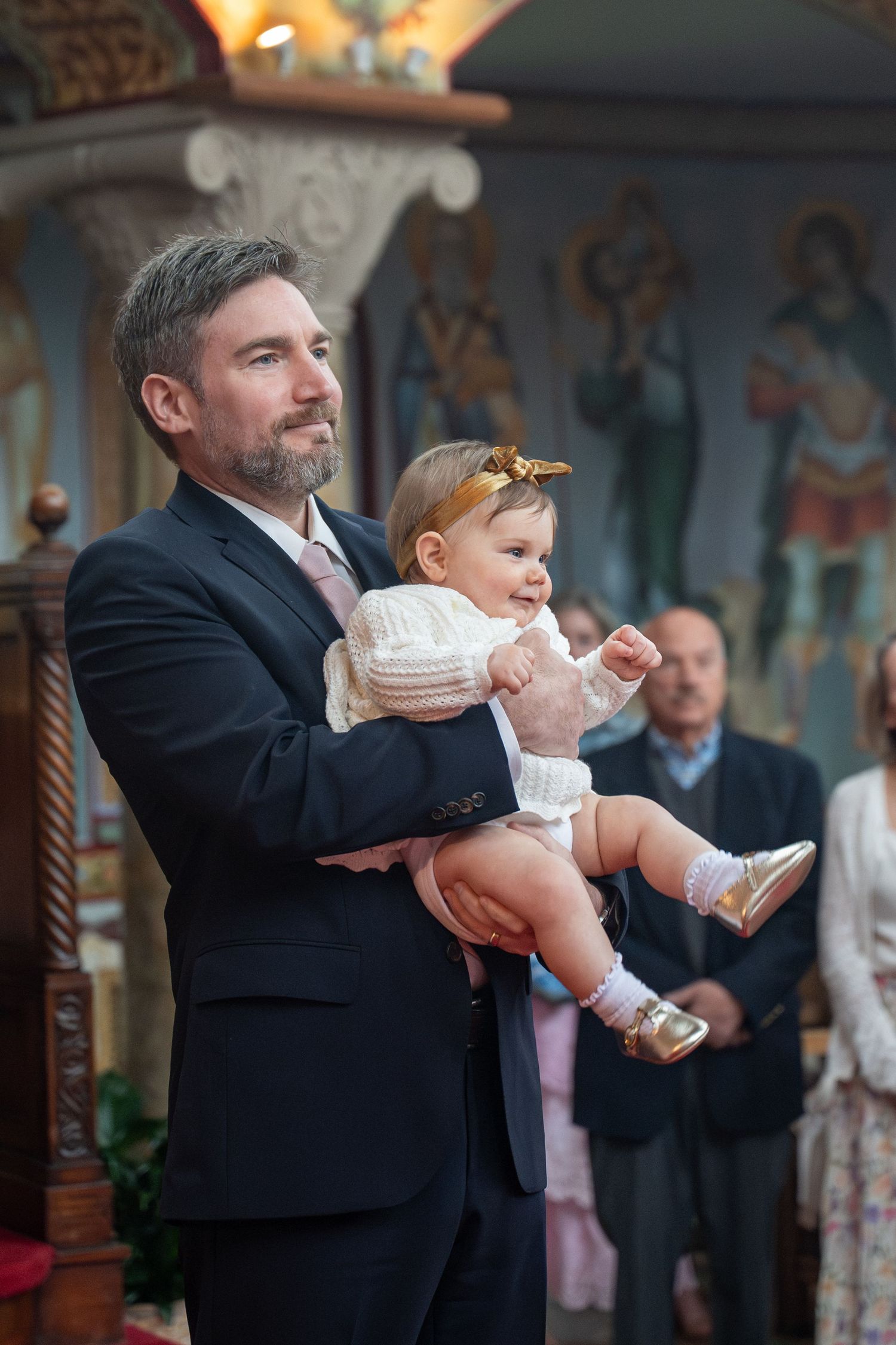 Man in suit holding baby girl in white dress during Orthodox church baptism ceremony.