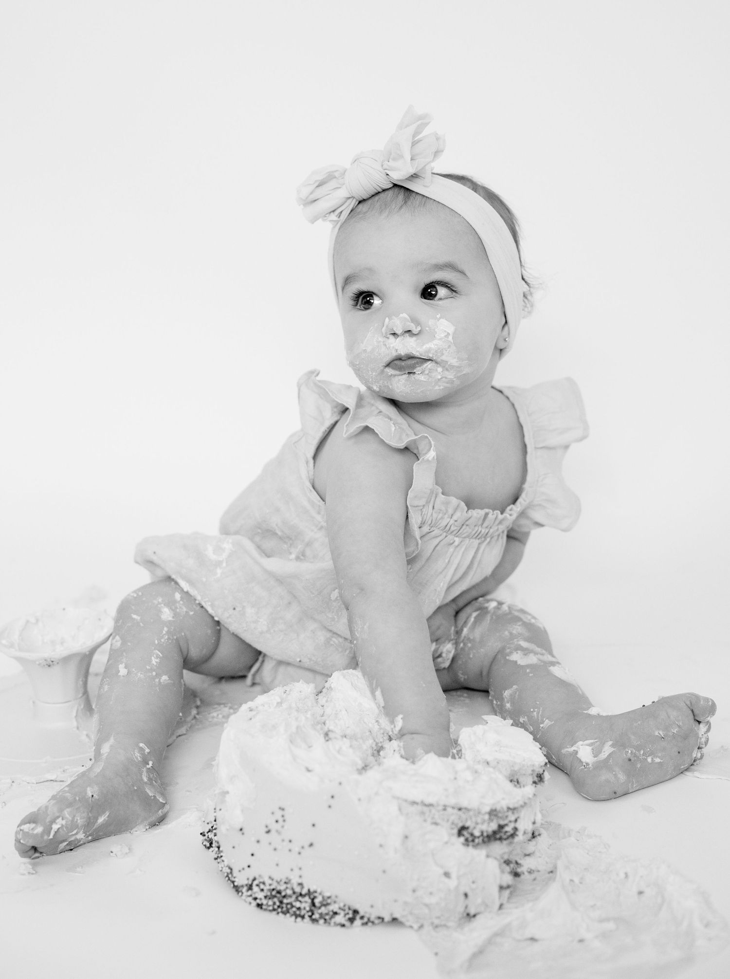 Black and white photo of baby in dress sitting with smashed cake.