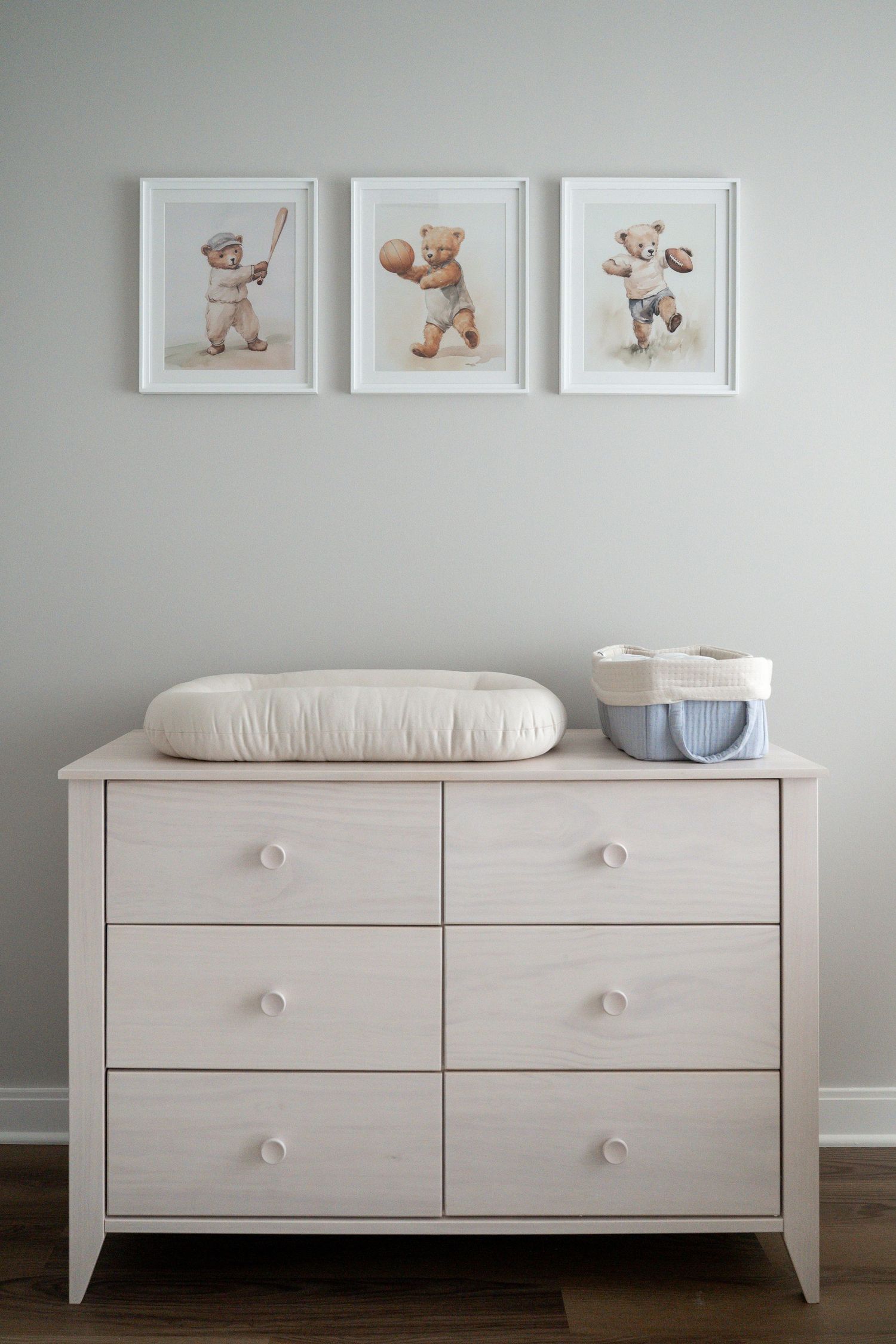 Light gray nursery dresser with changing pad and three framed teddy bear prints on wall above in minimalist style.