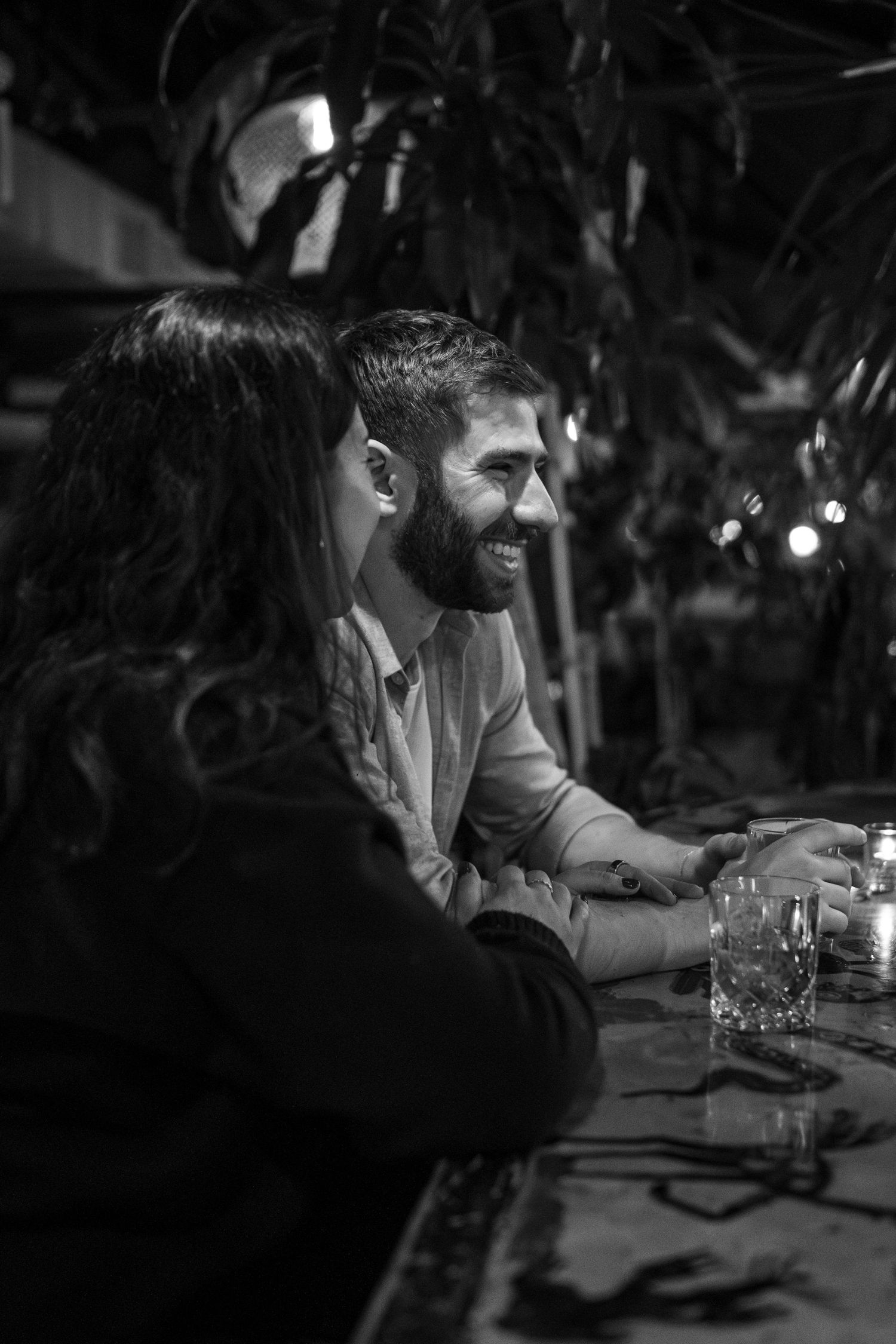 A couple shares drinks and laughter at a dimly lit bar in an intimate black and white moment.