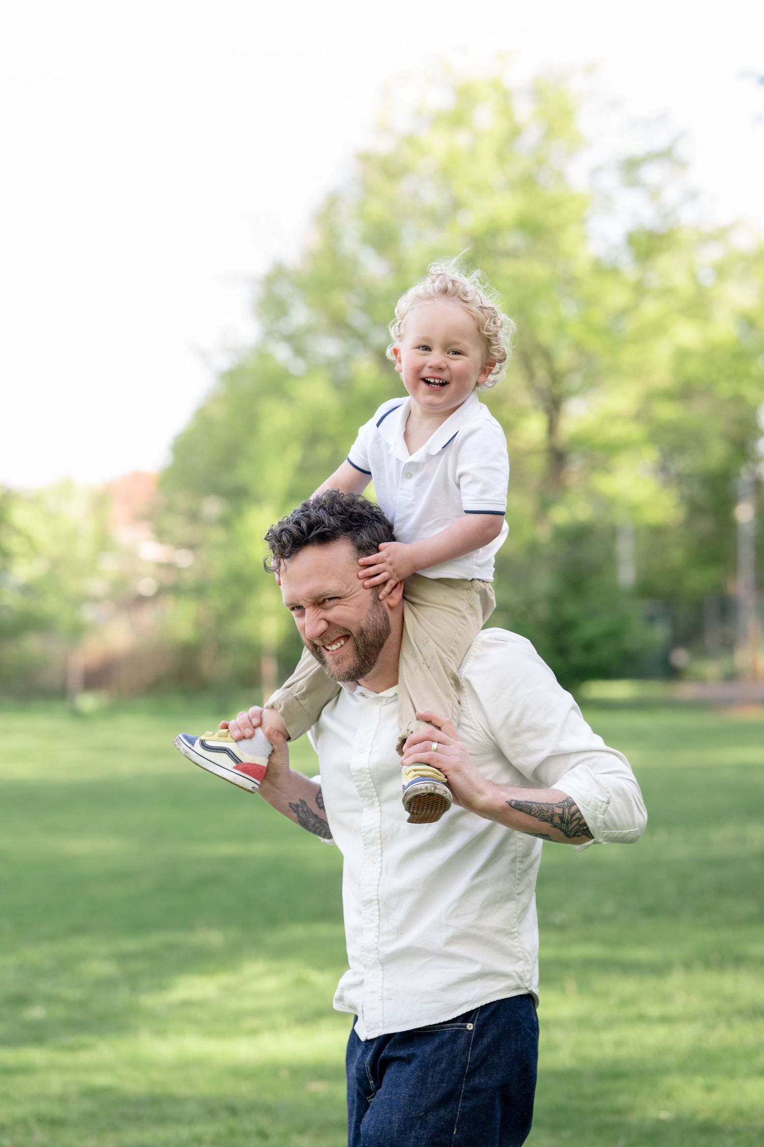 Happy father carrying smiling curly-haired toddler on shoulders in sunny green park.