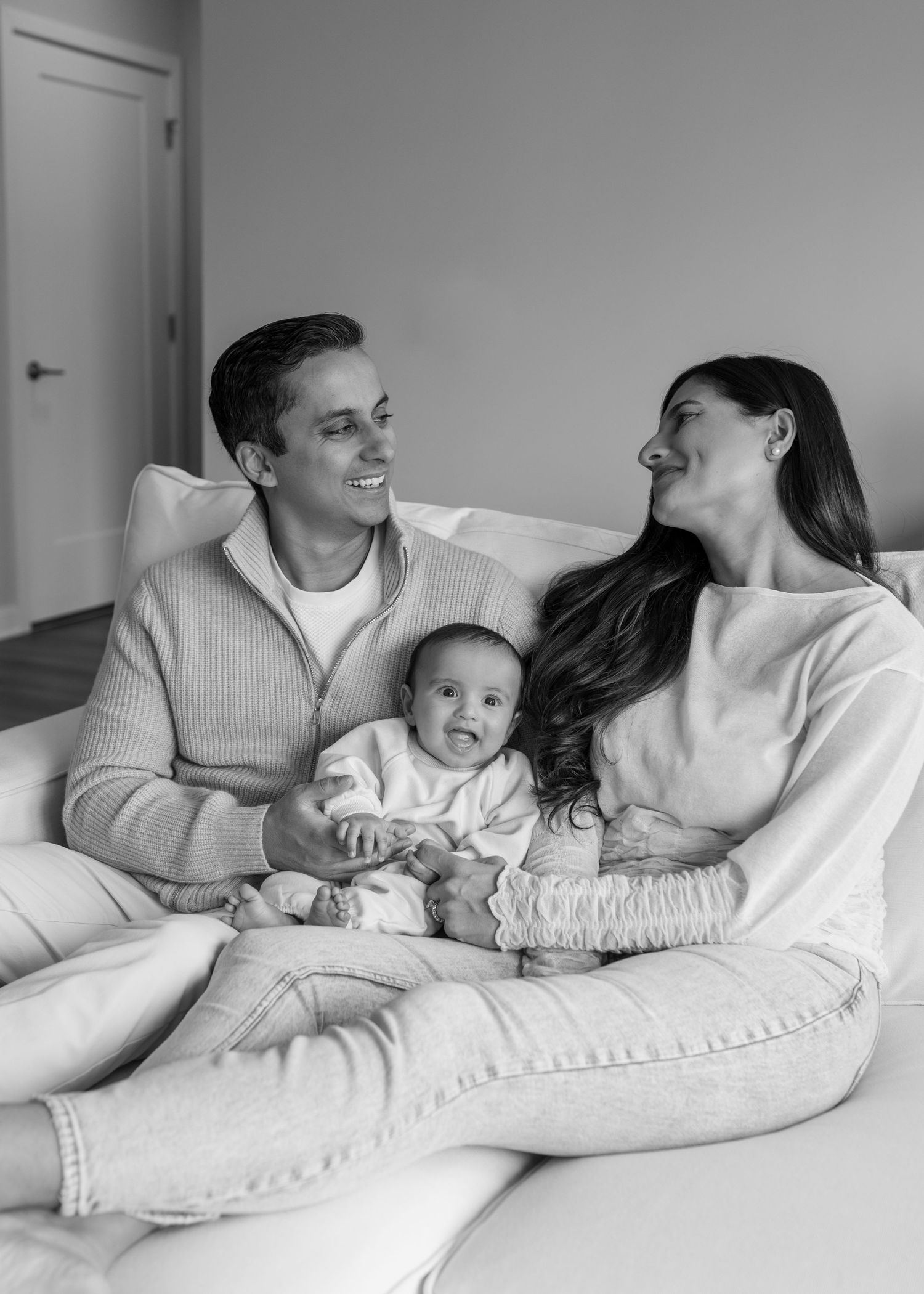 Black and white photo of happy parents sitting on couch with their cute baby between them.