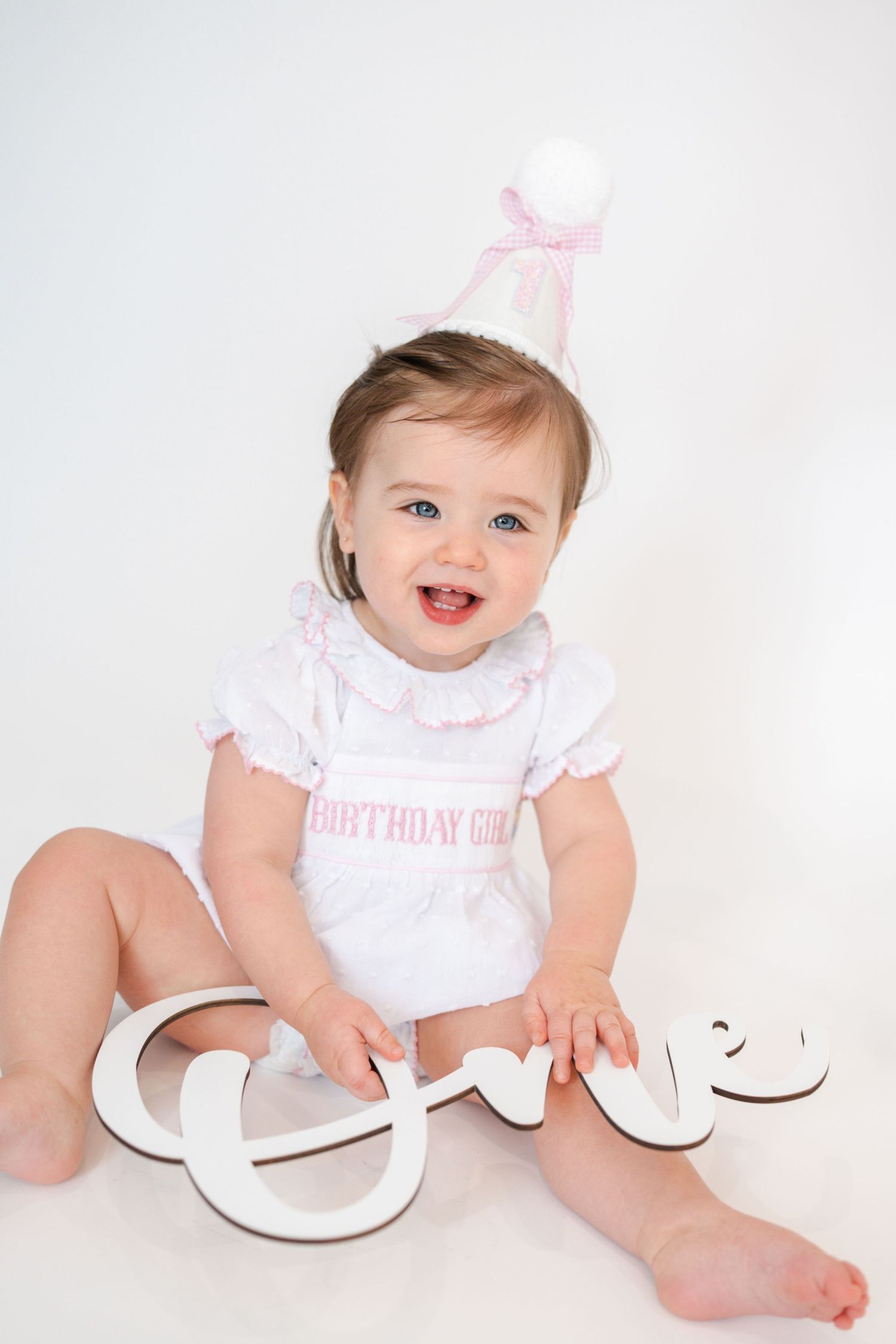 Baby in white dress sitting with wooden 'ONE' letters against white background.