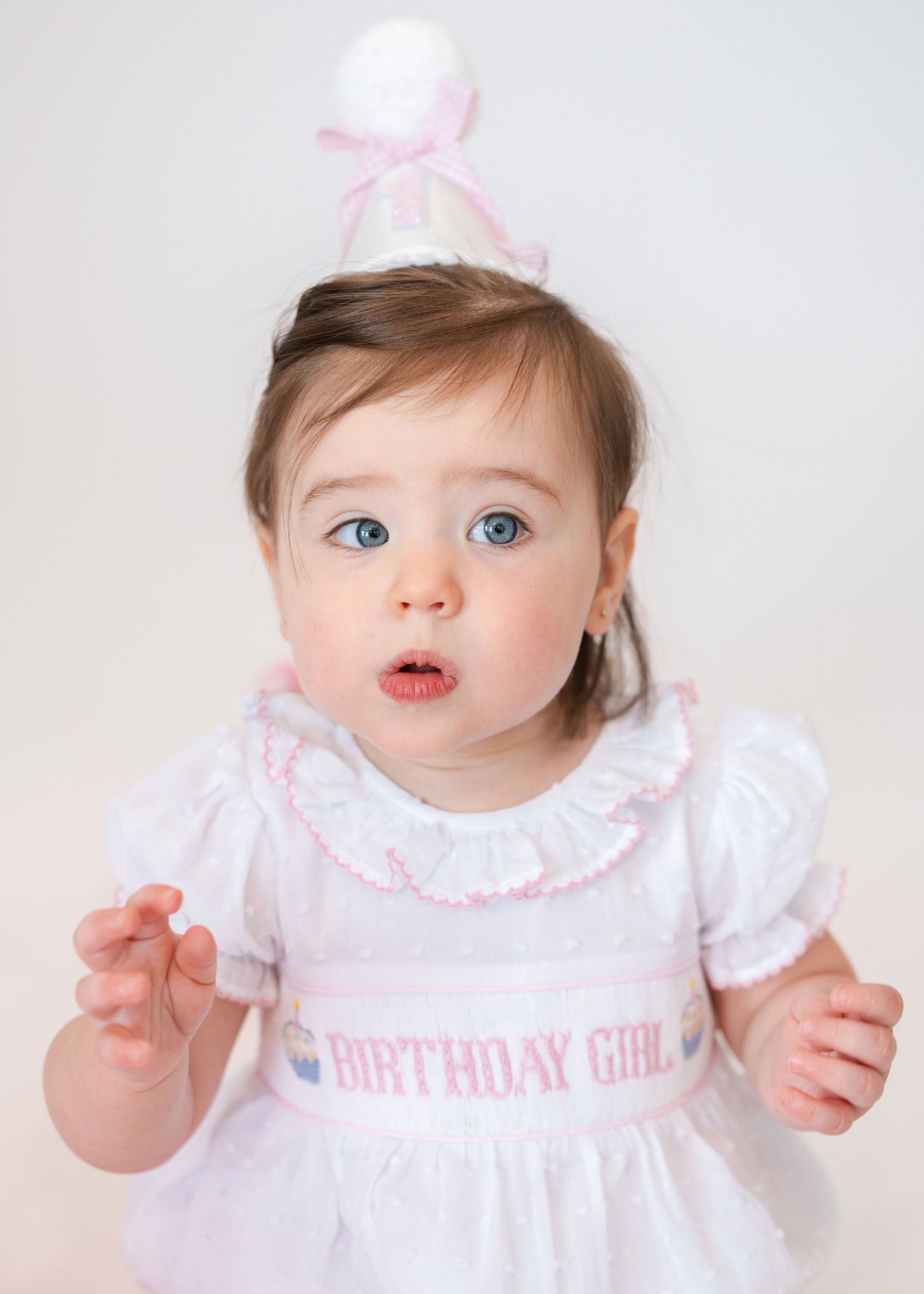 Baby in white dress with pink accents reaches out during photoshoot.