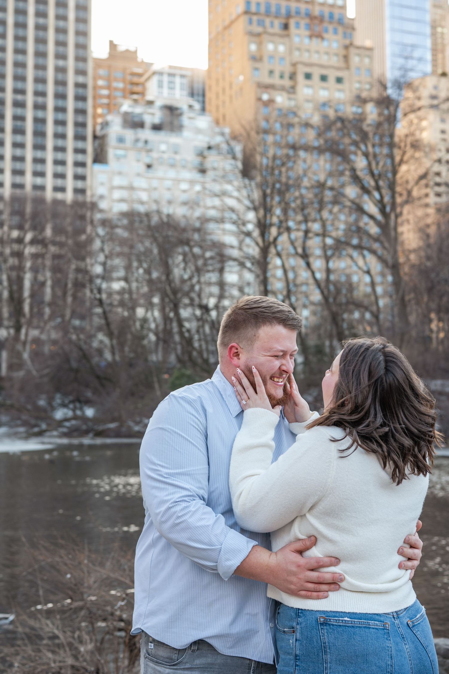 A couple sharing an emotional embrace by a lake with winter trees and urban buildings in the background.